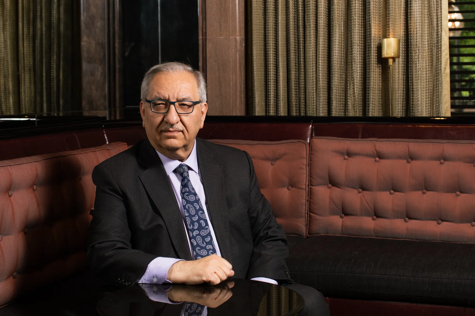 A middle-aged man with gray hair, wearing glasses, a black suit, a purple shirt, and a patterned tie, sitting on a pink cushioned bench in a sophisticated room with wood-paneled walls, dark curtains, and a gold wall lamp.