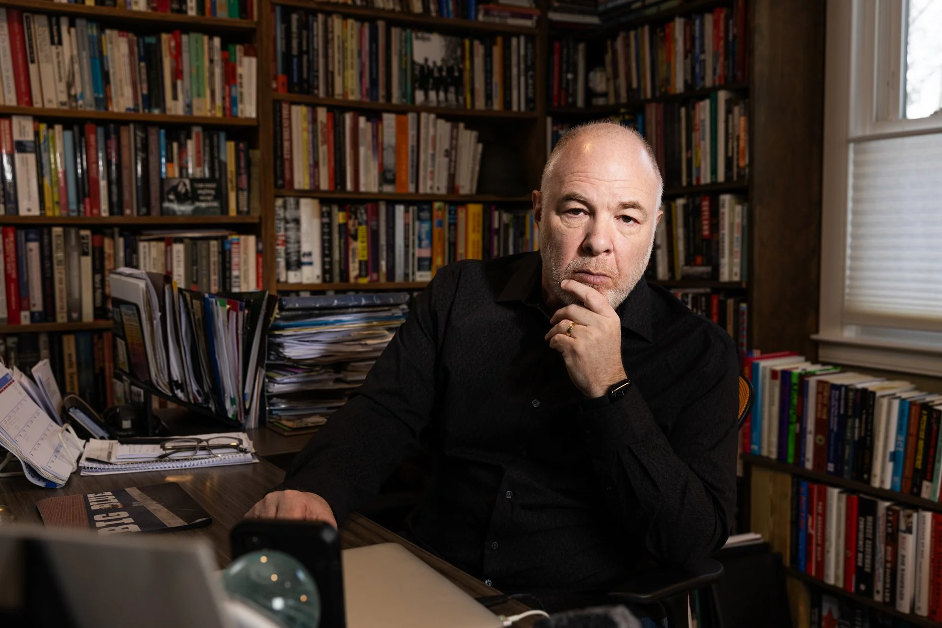 A middle-aged man with a bald head and gray beard, wearing a black shirt, seated at a cluttered desk in front of a large bookshelf filled with various books. He has a thoughtful expression, resting his chin on his hand, with one eye slightly squintin