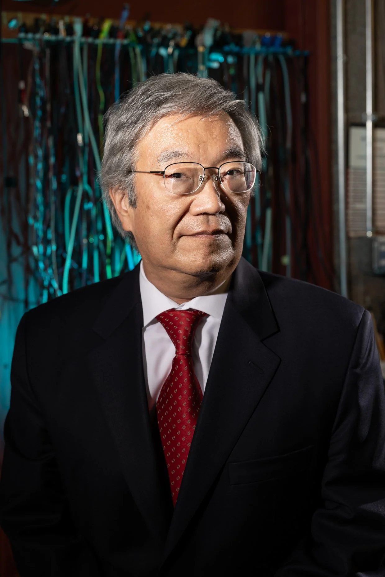 A middle-aged man with gray hair and glasses wearing a black suit, white shirt, and red tie, sitting indoors with a blurred background of cables or wires.