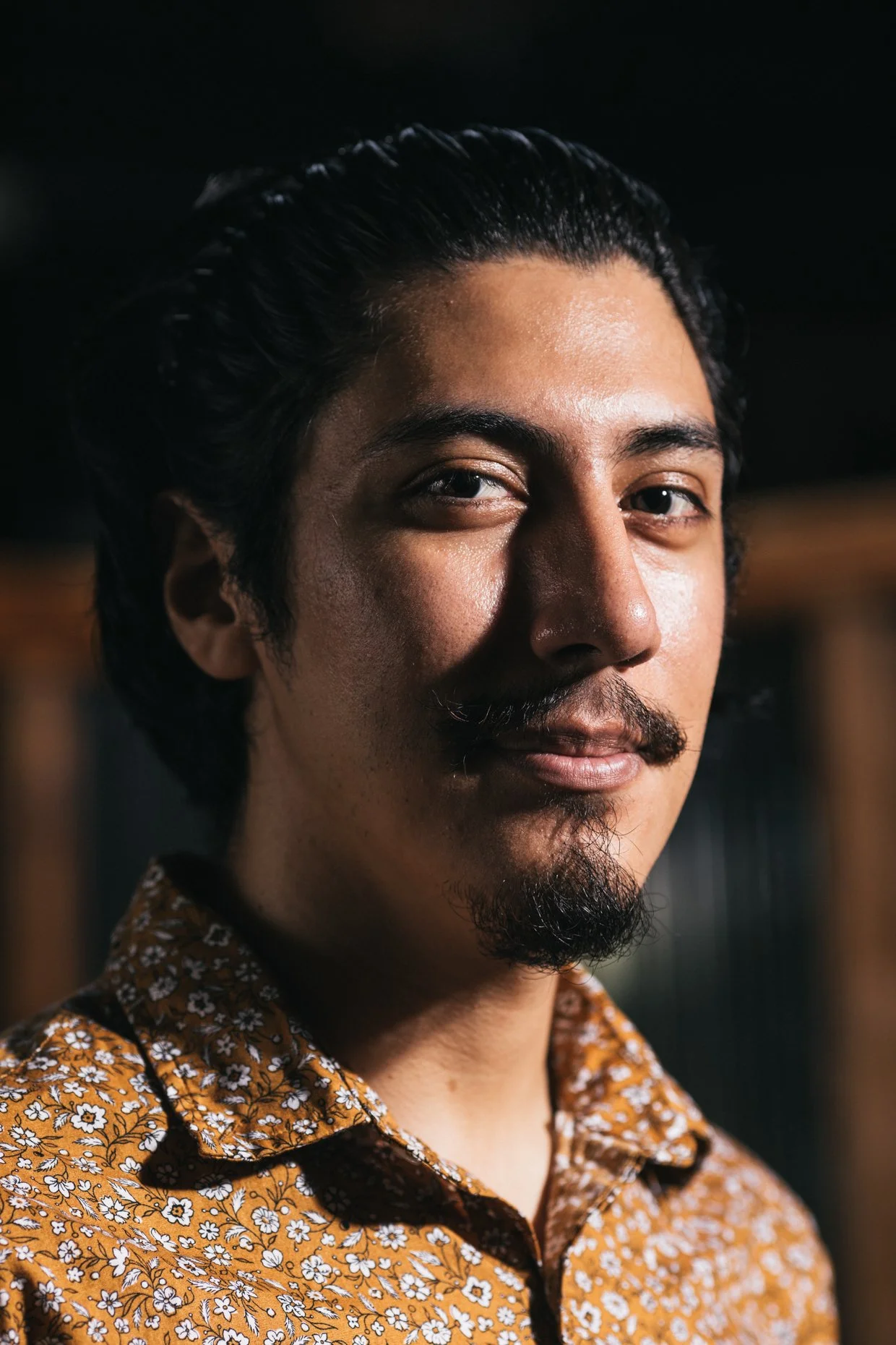 Close-up portrait of a man with dark hair, mustache, and goatee, wearing a patterned shirt, looking at the camera with a slight smile.