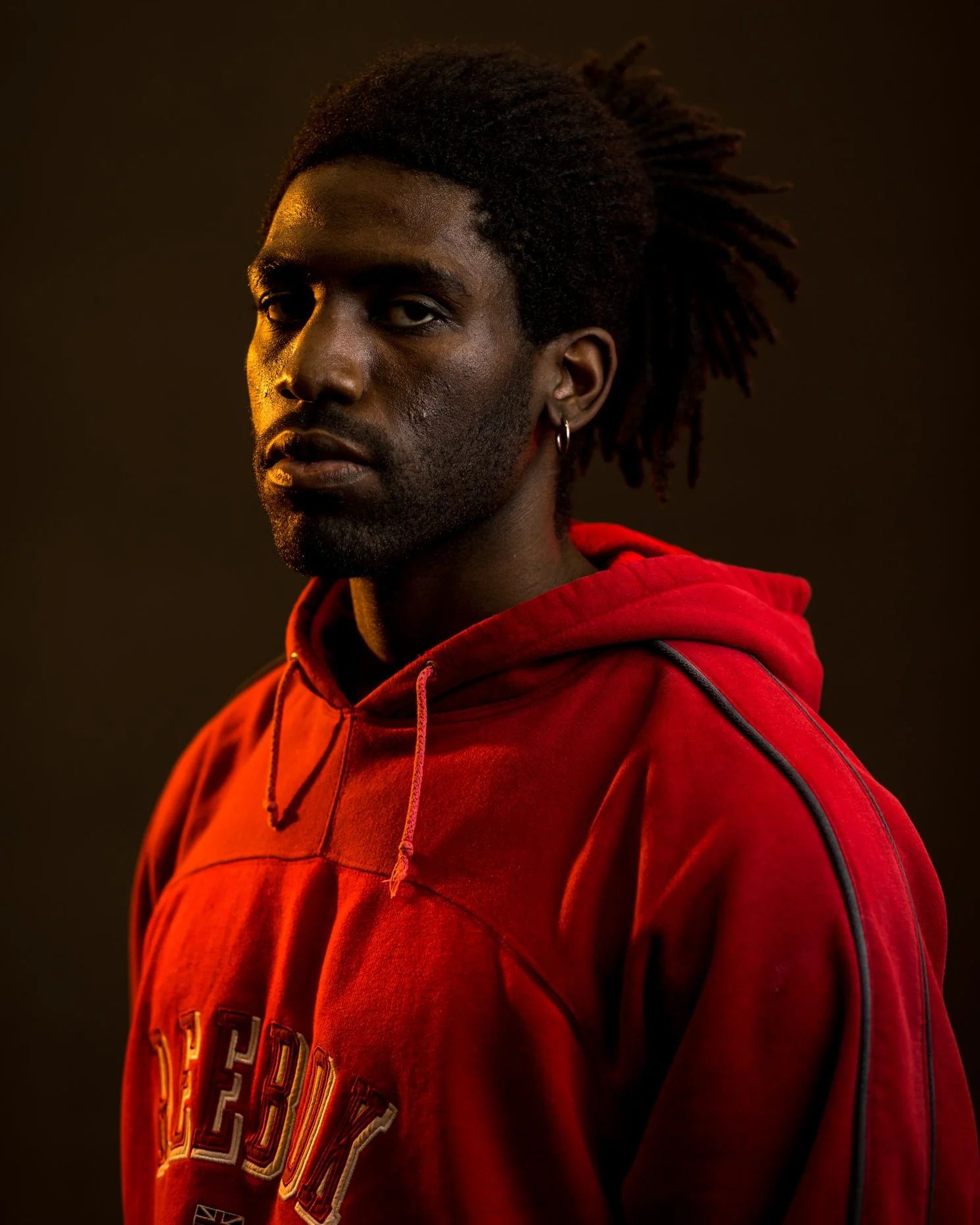 A young man with dark skin and dreadlocks wearing a red hoodie, looking to the side with a serious expression against a dark background.