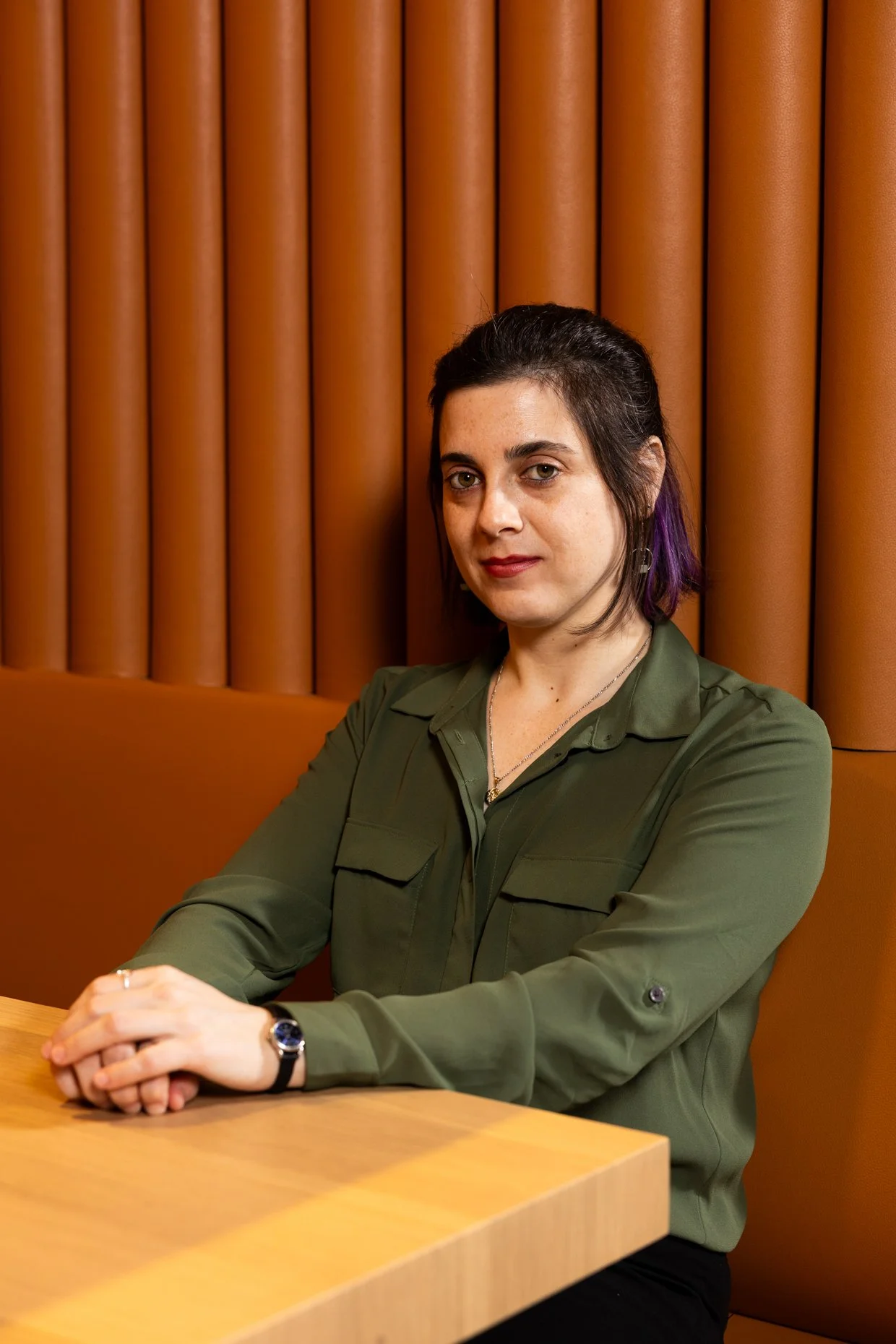 A woman with short dark hair and purple streaks, wearing a green button-up shirt, sitting at a wooden table against a background of orange vertical panels.