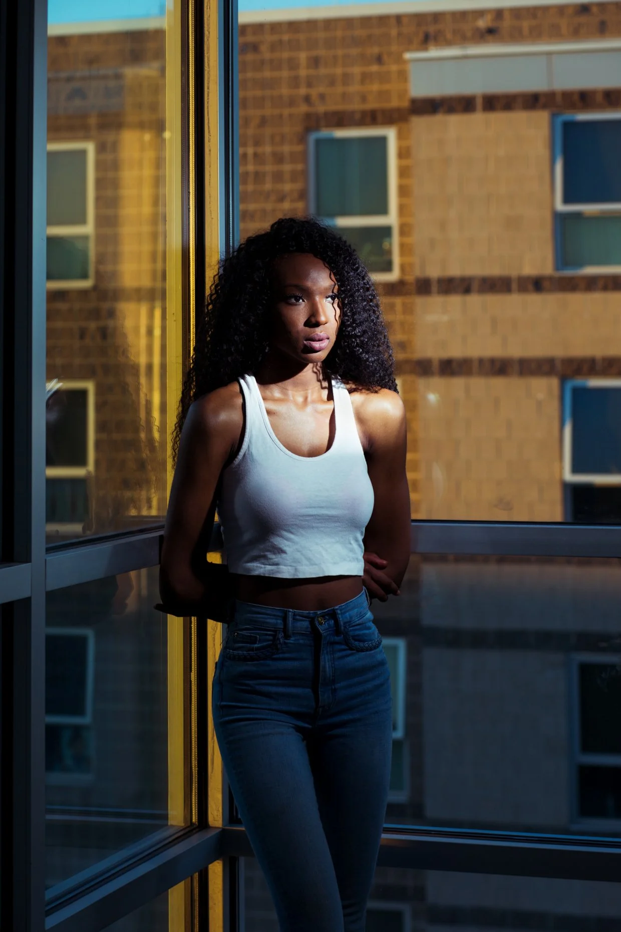 A young woman with curly hair and dark skin, wearing a white tank top and blue jeans, standing by a large window at sunset, with a brick building visible outside.