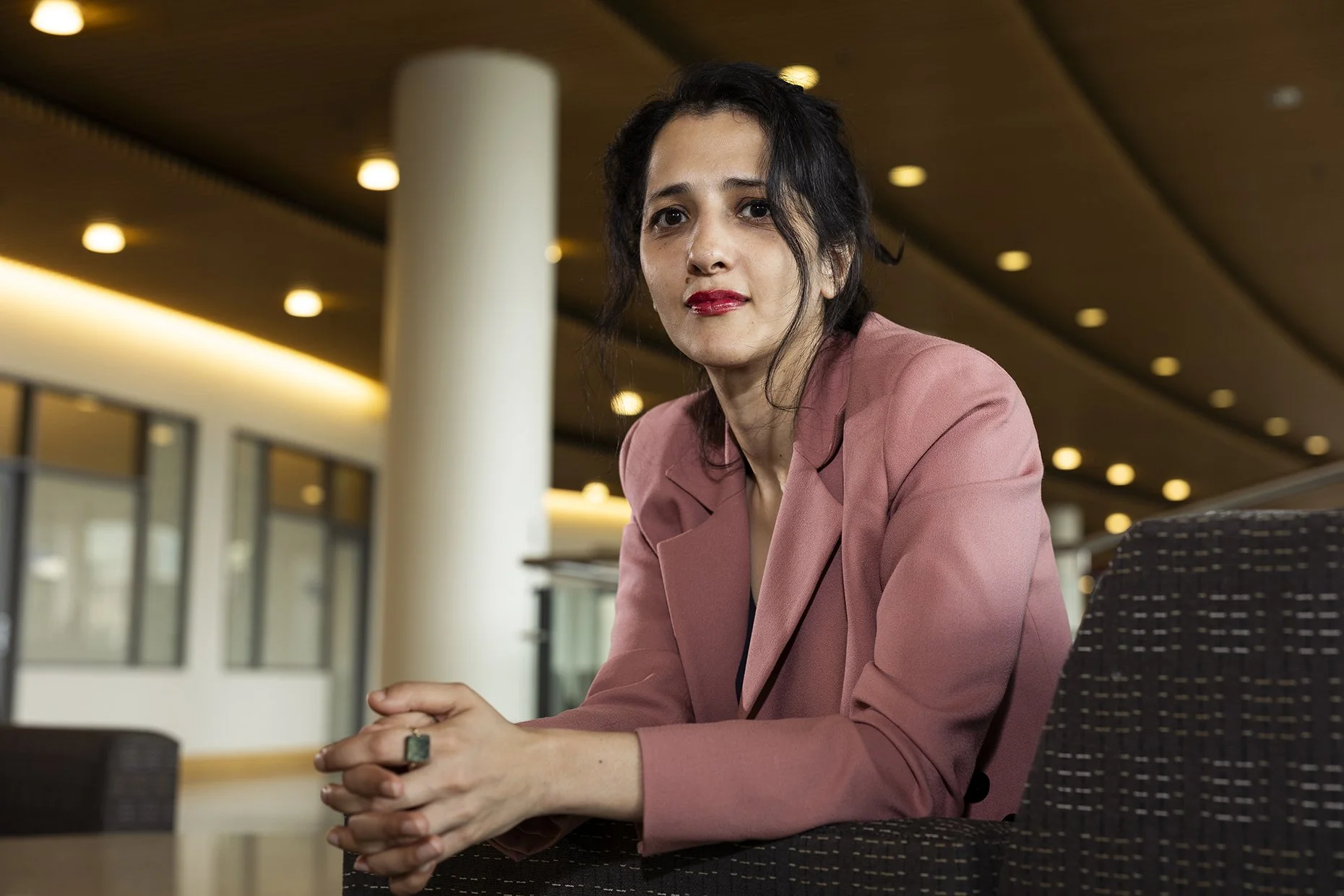 A woman with dark hair and red lipstick sitting at a table in a modern indoor space with warm lighting. She is wearing a pink blazer and has a large ring on her finger. She is looking directly at the camera with a slight smile, her hands clasped in f