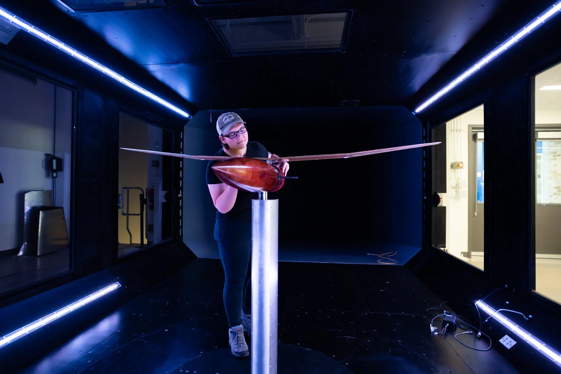 A person with glasses and a gray cap examines a large, elongated, shiny wooden object mounted on a metal pedestal in a dark room with blue strip lights along the ceiling.