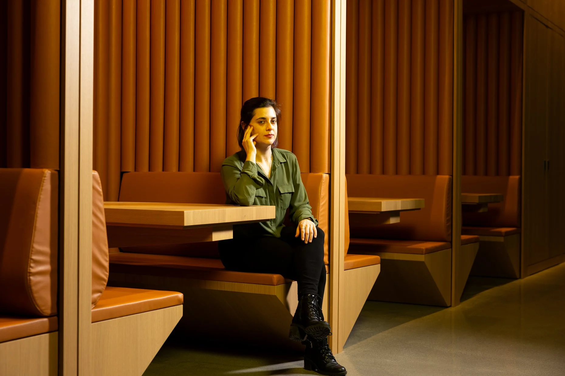 A woman with dark hair sitting in a booth at a restaurant or cafe, wearing a green shirt and black pants, with her hand resting on her face. The booth has wooden panels and orange cushions.