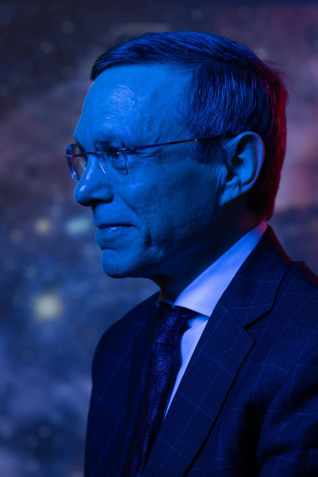 Side profile of a man with short dark hair and glasses wearing a dark checked suit, white shirt, and patterned tie, illuminated with blue lighting.