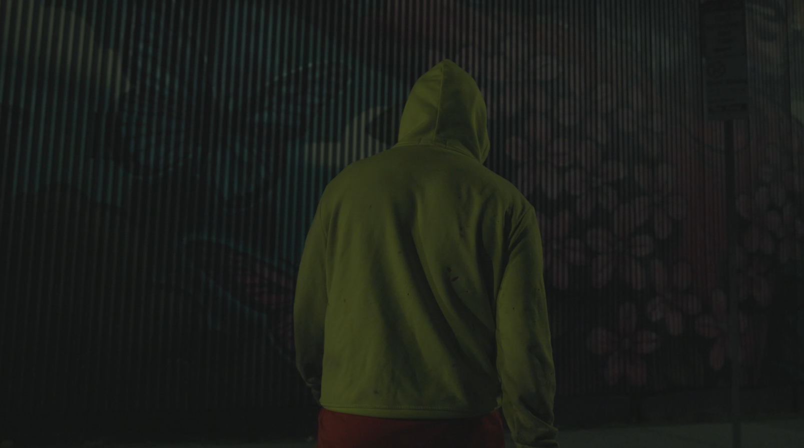 A person wearing a yellow hoodie stands with their back to the camera in front of a large, illuminated LED display with colorful patterns.