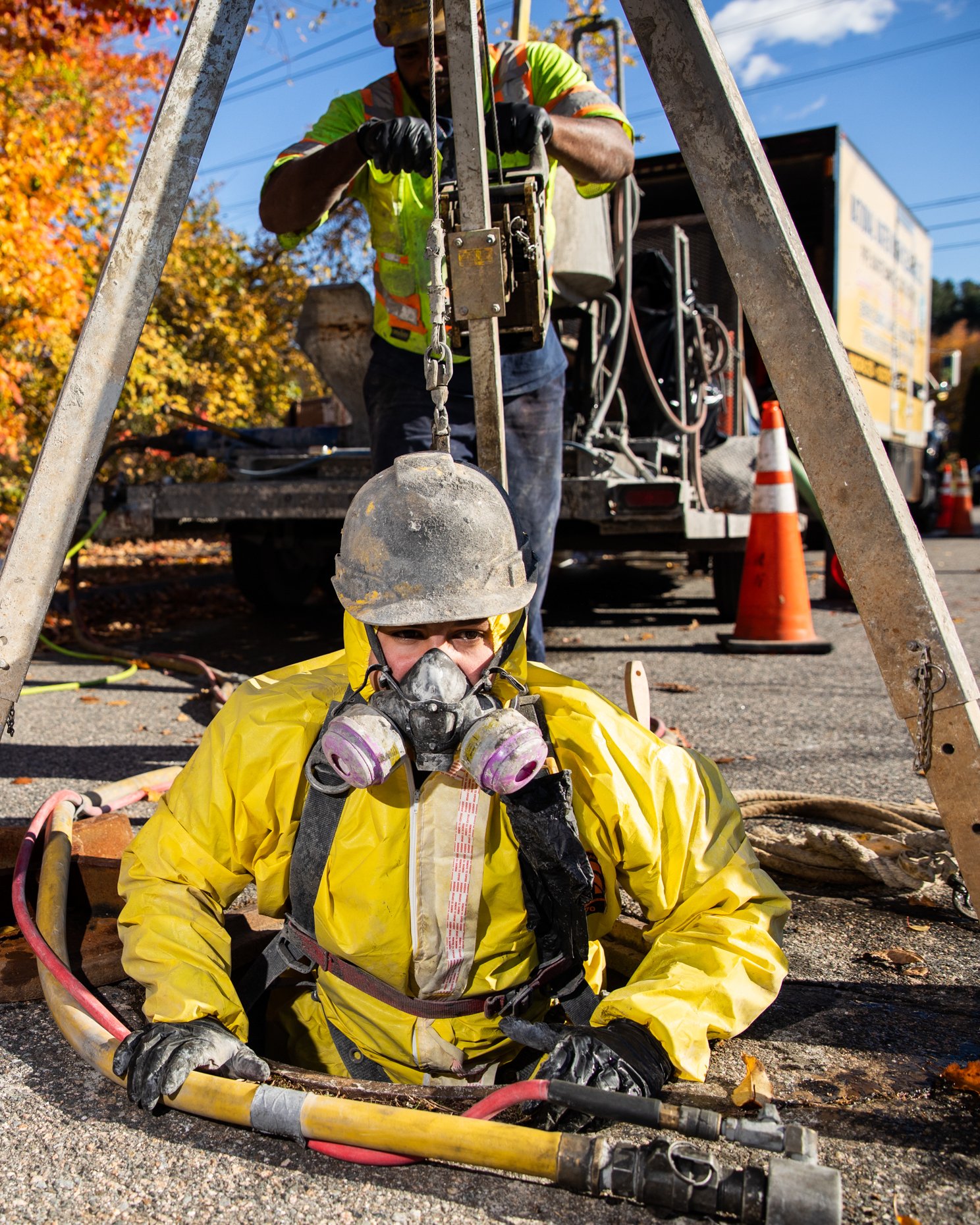 A worker in a yellow rain suit and protective helmet sitting on the ground with a respirator mask. Behind them, a worker in green safety vest and helmet is working on electrical equipment. The scene appears to be outdoors with fall foliage and traffic cones visible.