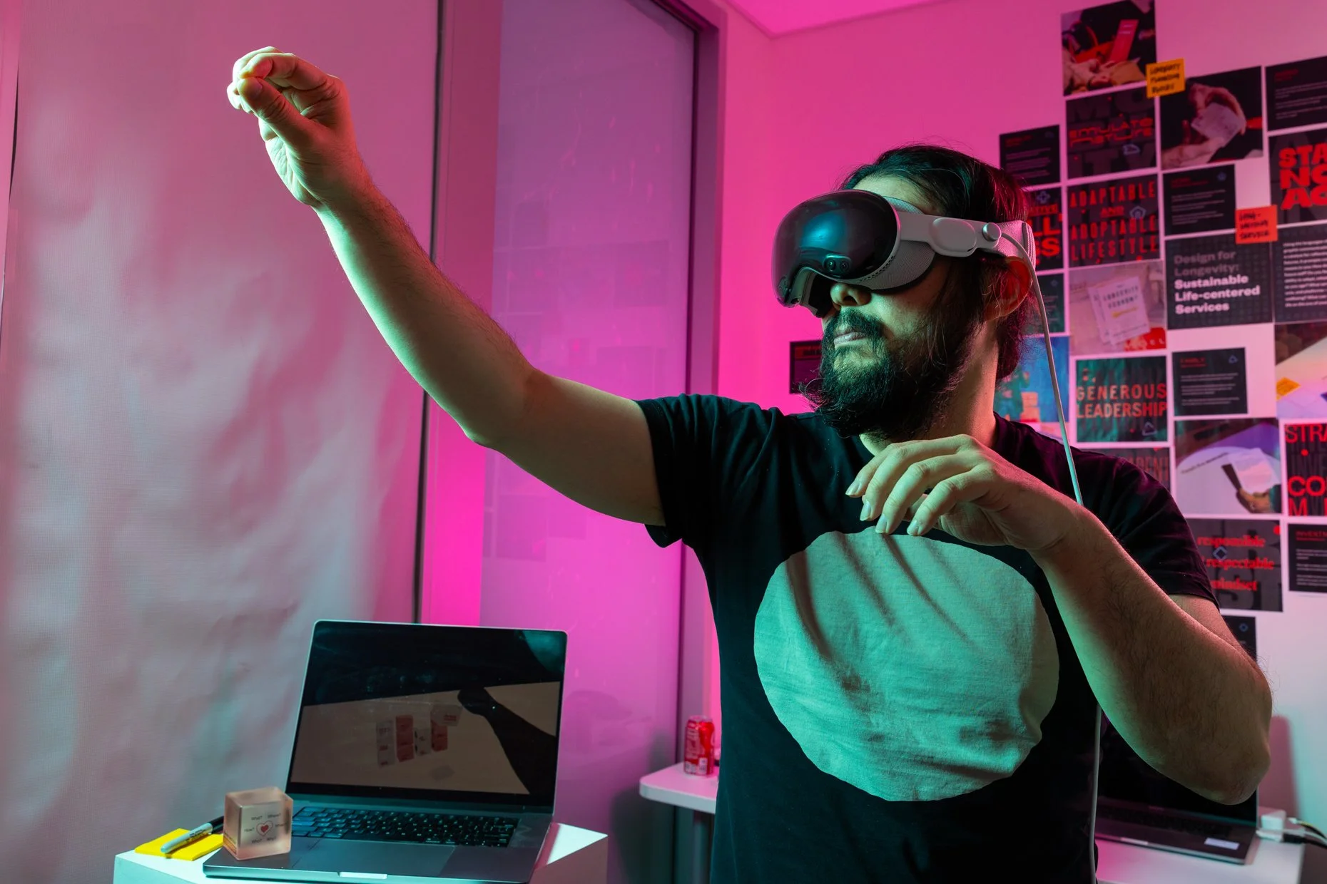 A man with a beard wearing a virtual reality headset, extending his right arm as if interacting with a virtual environment in a room with pink and purple lighting, posters on the wall, and a laptop and desk accessories on a table.