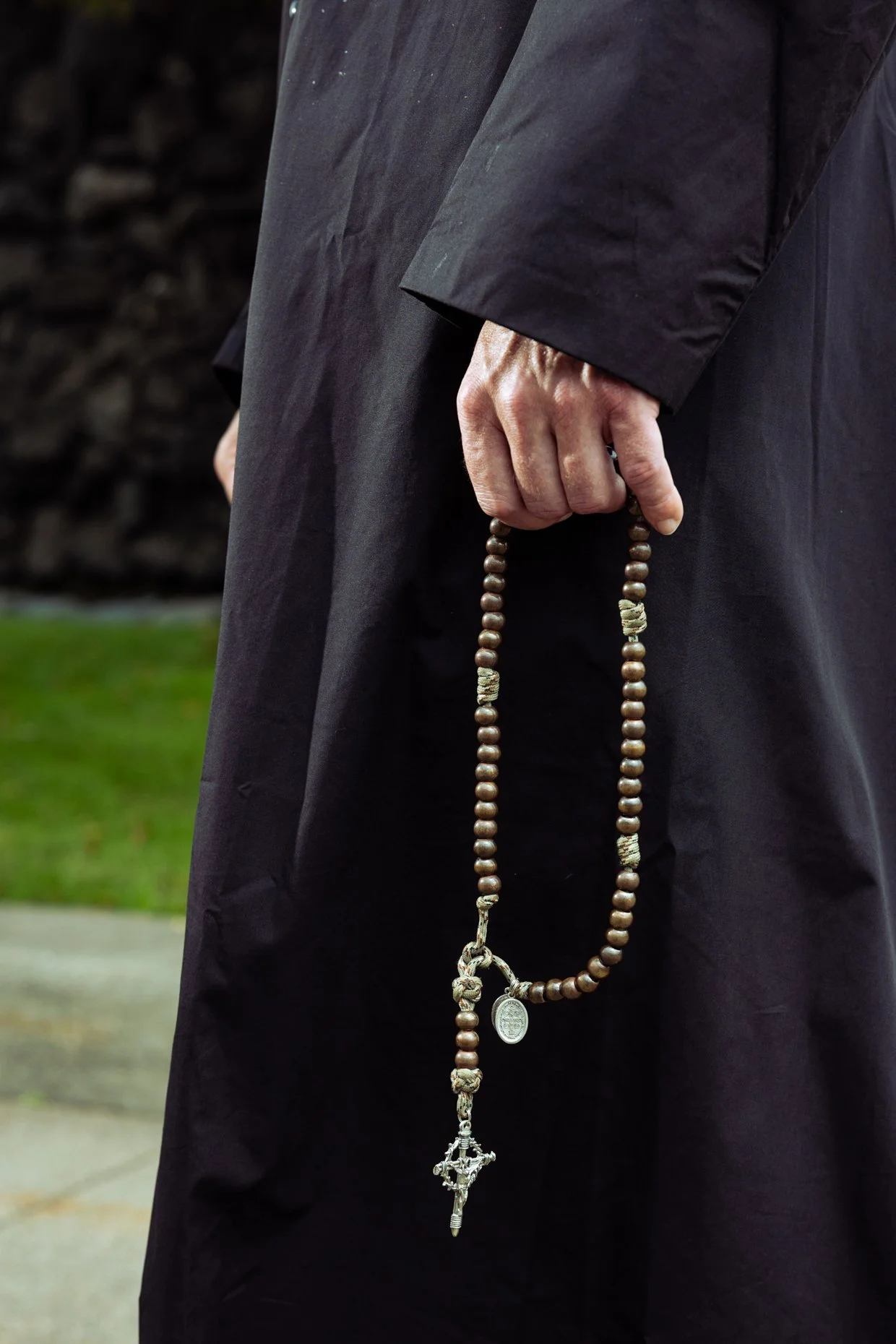 Close-up of a person wearing a black coat holding a rosary with wooden beads and a crucifix, outdoors with a grassy background.