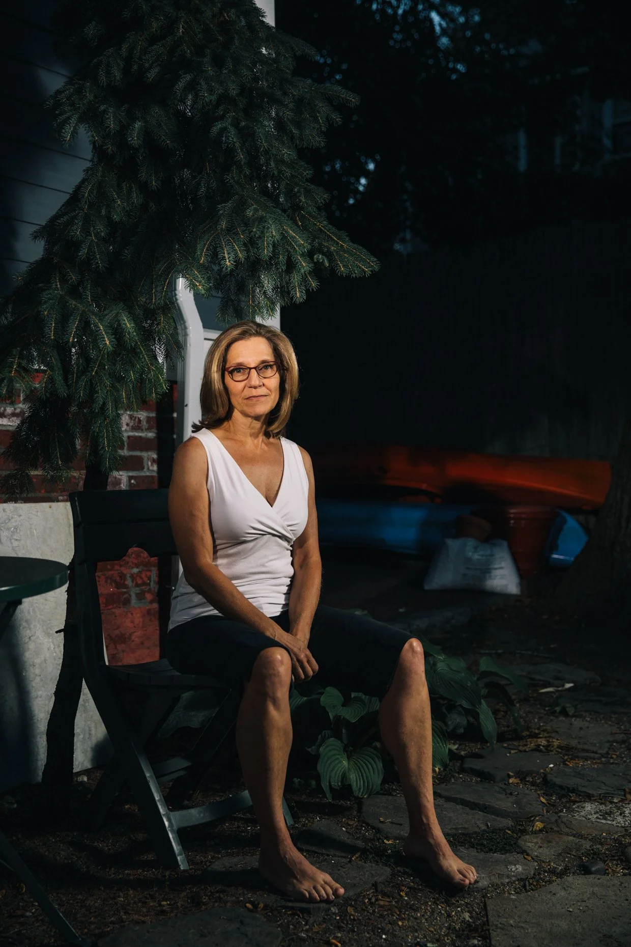 A woman sitting outdoors at night under a large pine tree. She is wearing glasses, a white sleeveless top, and black shorts. Her feet are bare, and she is seated on a black folding chair, with a small green table to her left. There are plants, a brick wall, and a red boat in the background.