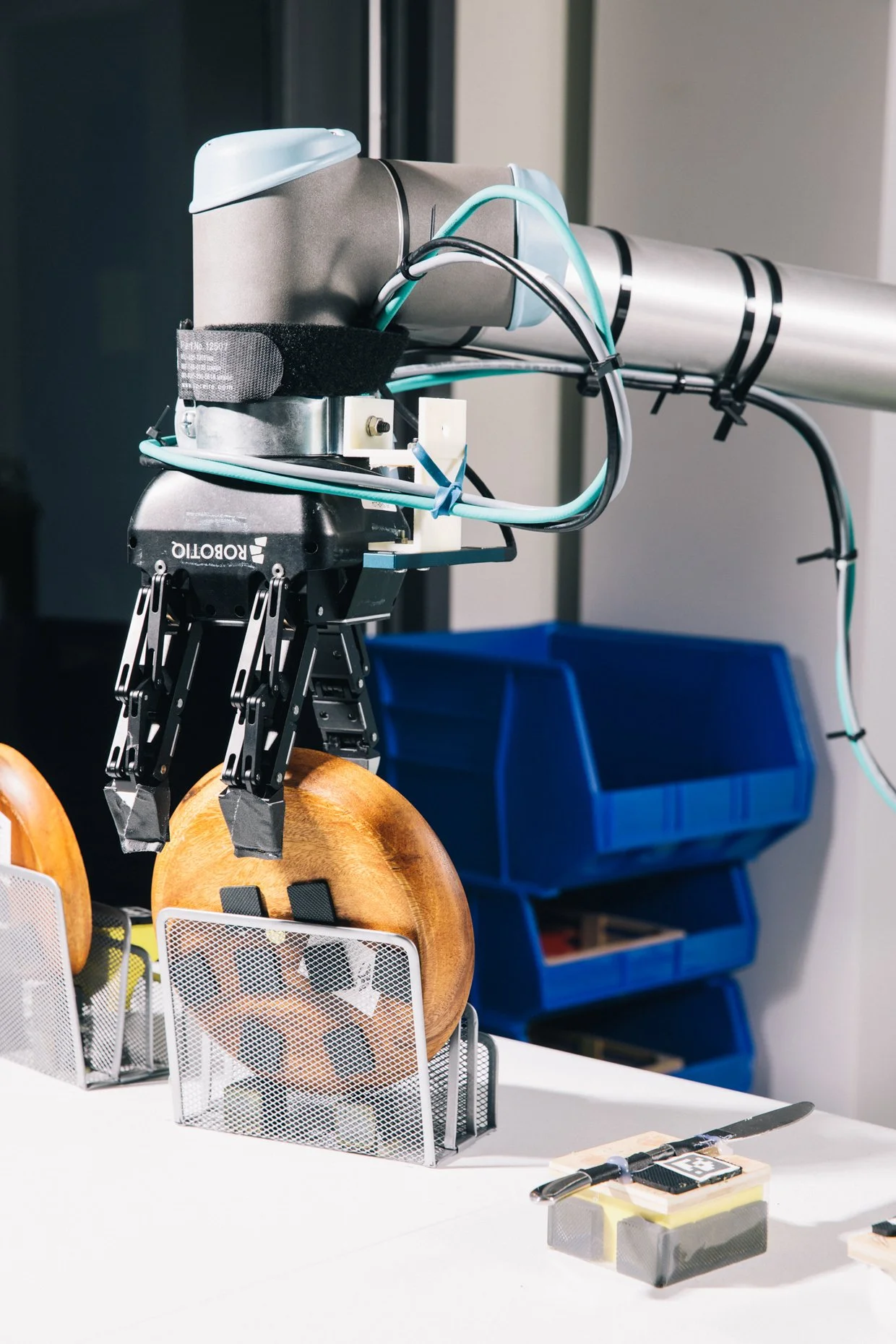 A robotic arm on a white table with dental tools and a small white basket holding two wooden objects. In the background, there are blue storage bins and a black wall.