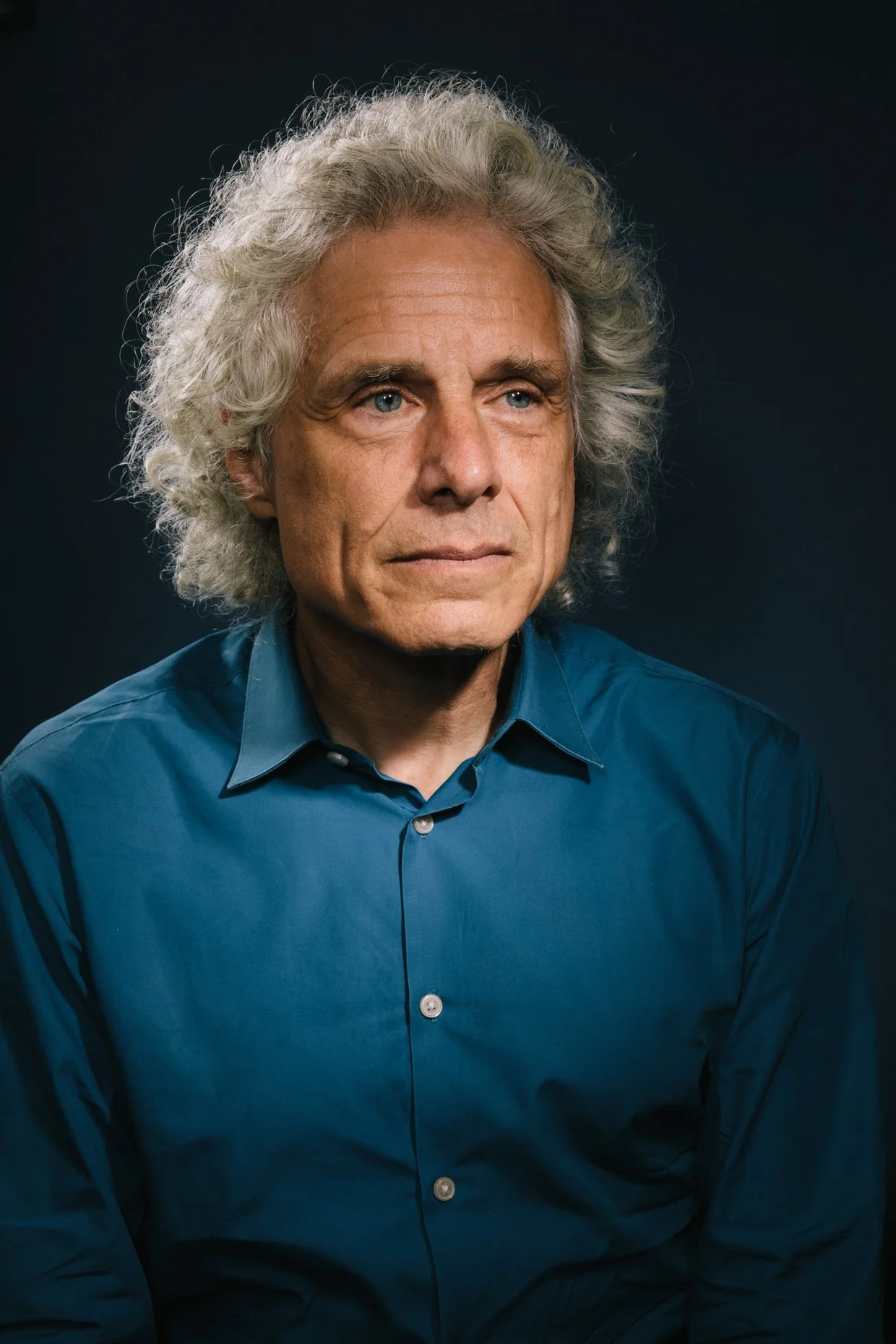 Portrait of an older man with curly gray hair, blue eyes, wearing a blue button-up shirt against a dark background.