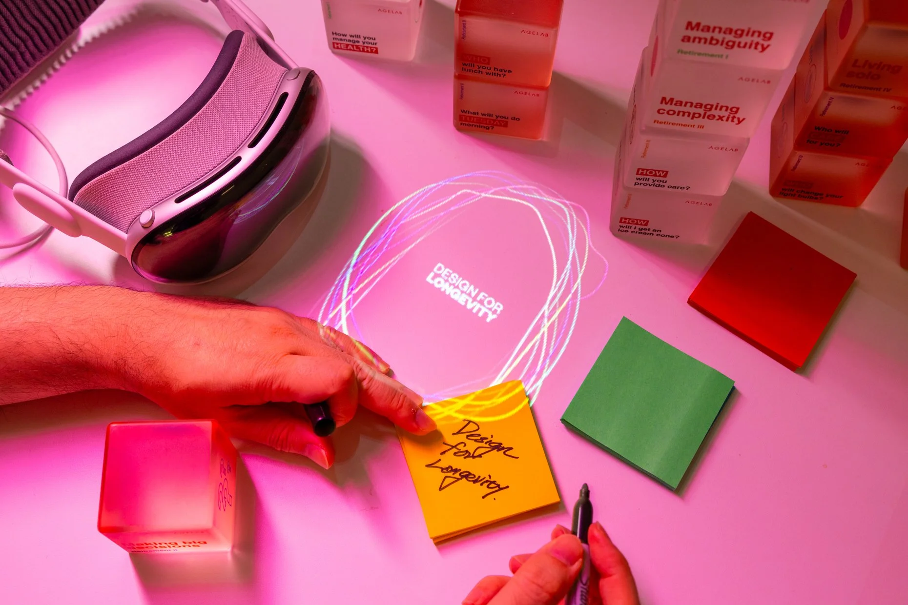 A table with colorful sticky notes, blocks with discussion questions, a VR headset, and a person writing on sticky notes under pink lighting, with the words "Design for Longevity" projected on the table.
