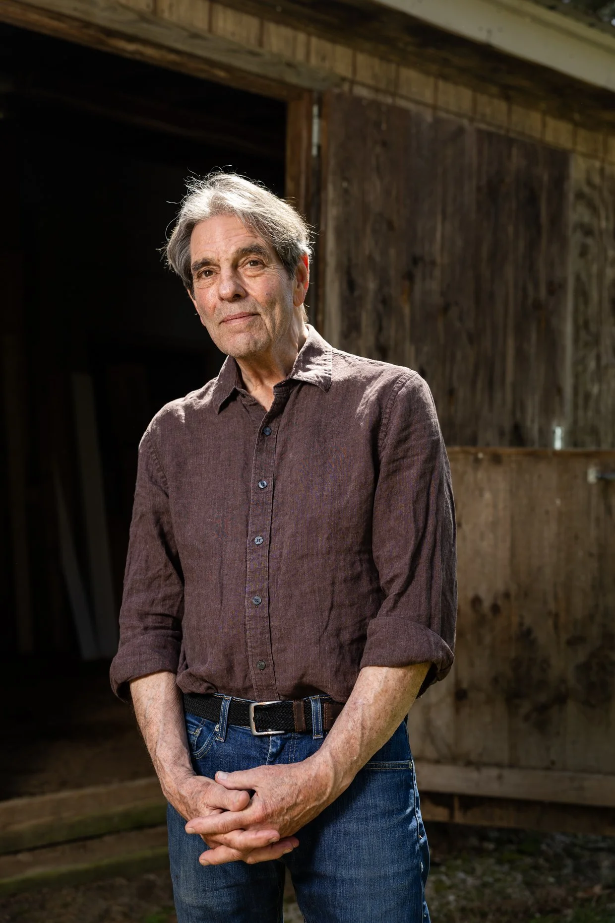 A mature man with gray hair and a slight smile standing outdoors in front of a wooden barn or shed, wearing a brown button-up shirt and jeans.