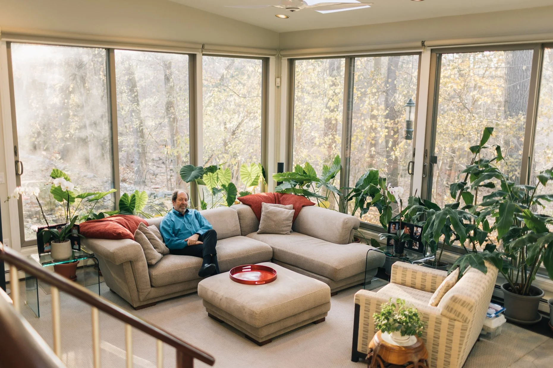 A man sitting on a beige sectional sofa in a sunlit living room with large windows and surrounded by green houseplants, a small table with a framed family photo, and two patterned armchairs in the foreground.