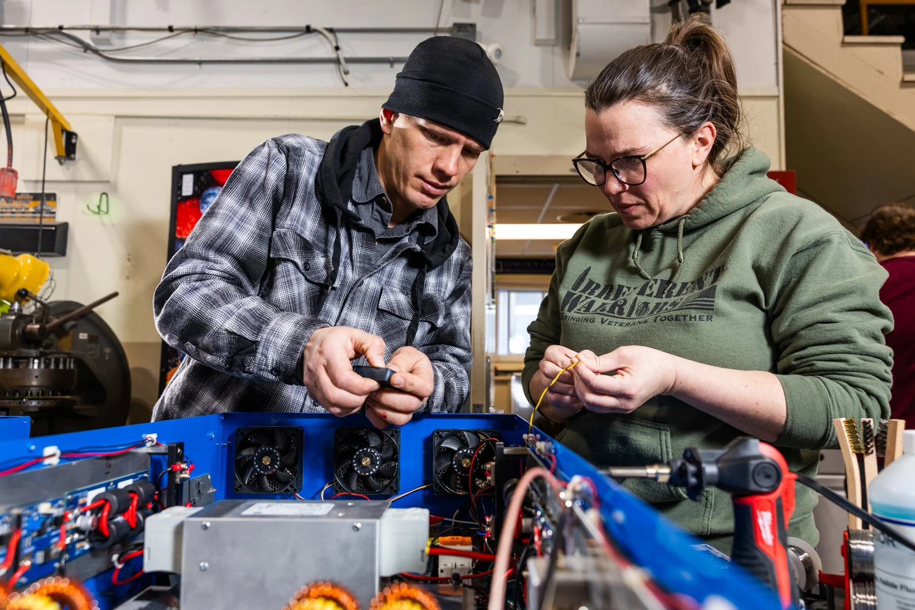Two individuals working on an electronic project in a workshop. One person is wearing a black beanie and a plaid shirt, and the other is wearing glasses and a green hoodie. They are focused on assembling or repairing electronic components inside a blue casing with various wires and electronic parts visible.