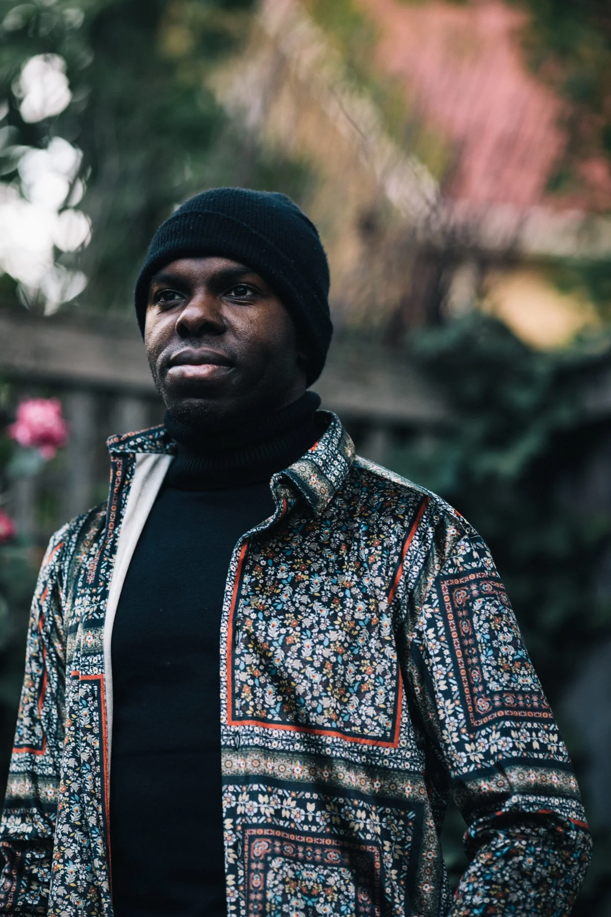 A man wearing a black knit hat and a multicolored floral patterned shirt over a black turtleneck, standing outdoors with blurred foliage in the background.