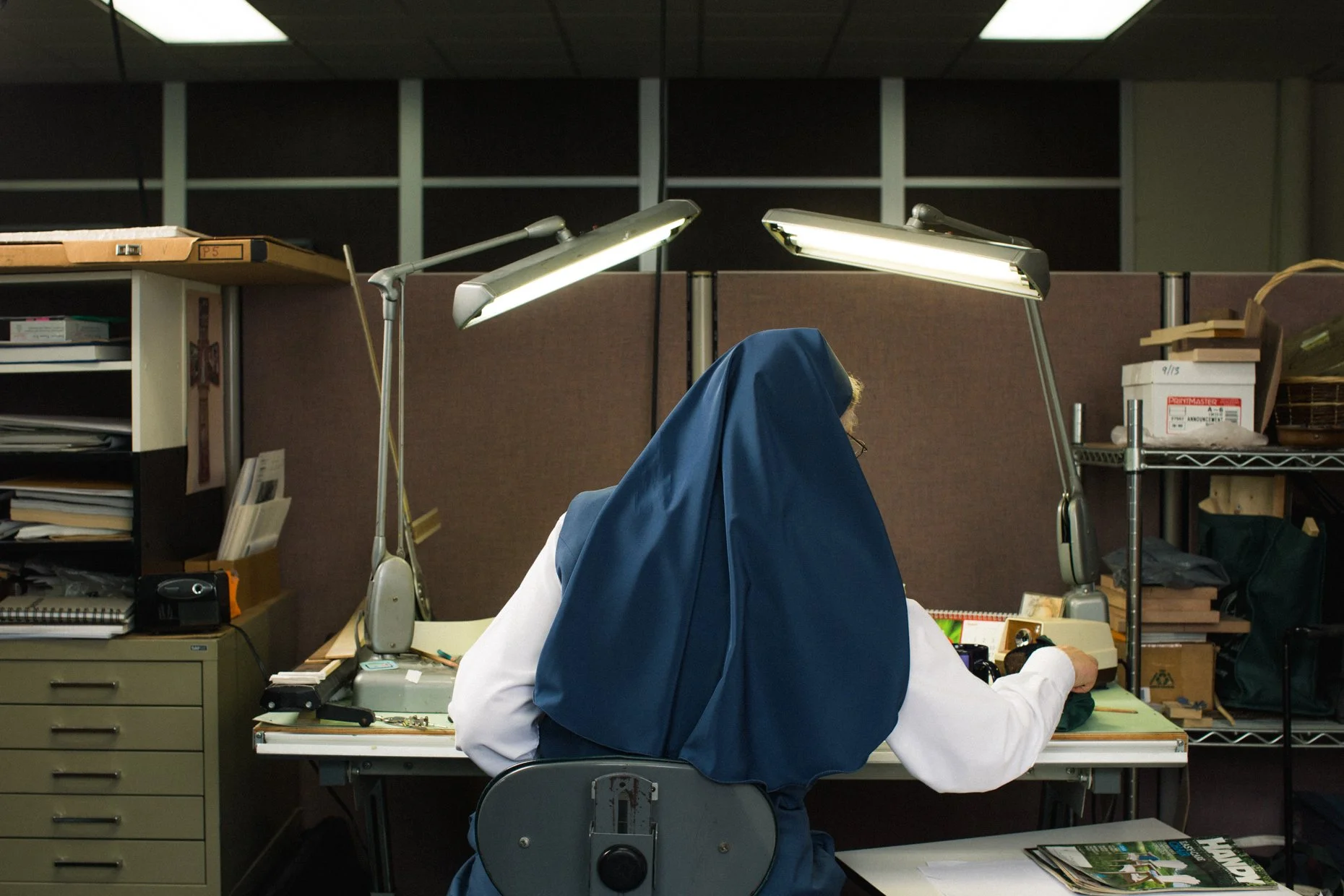 A person seated at a desk in an office cubicle, wearing a white shirt and a blue hood that covers their head. The desk has overlapping green folders and assorted office supplies, illuminated by two desk lamps. The background shows office shelves with files and boxes, and a dark ceiling with a grid pattern.