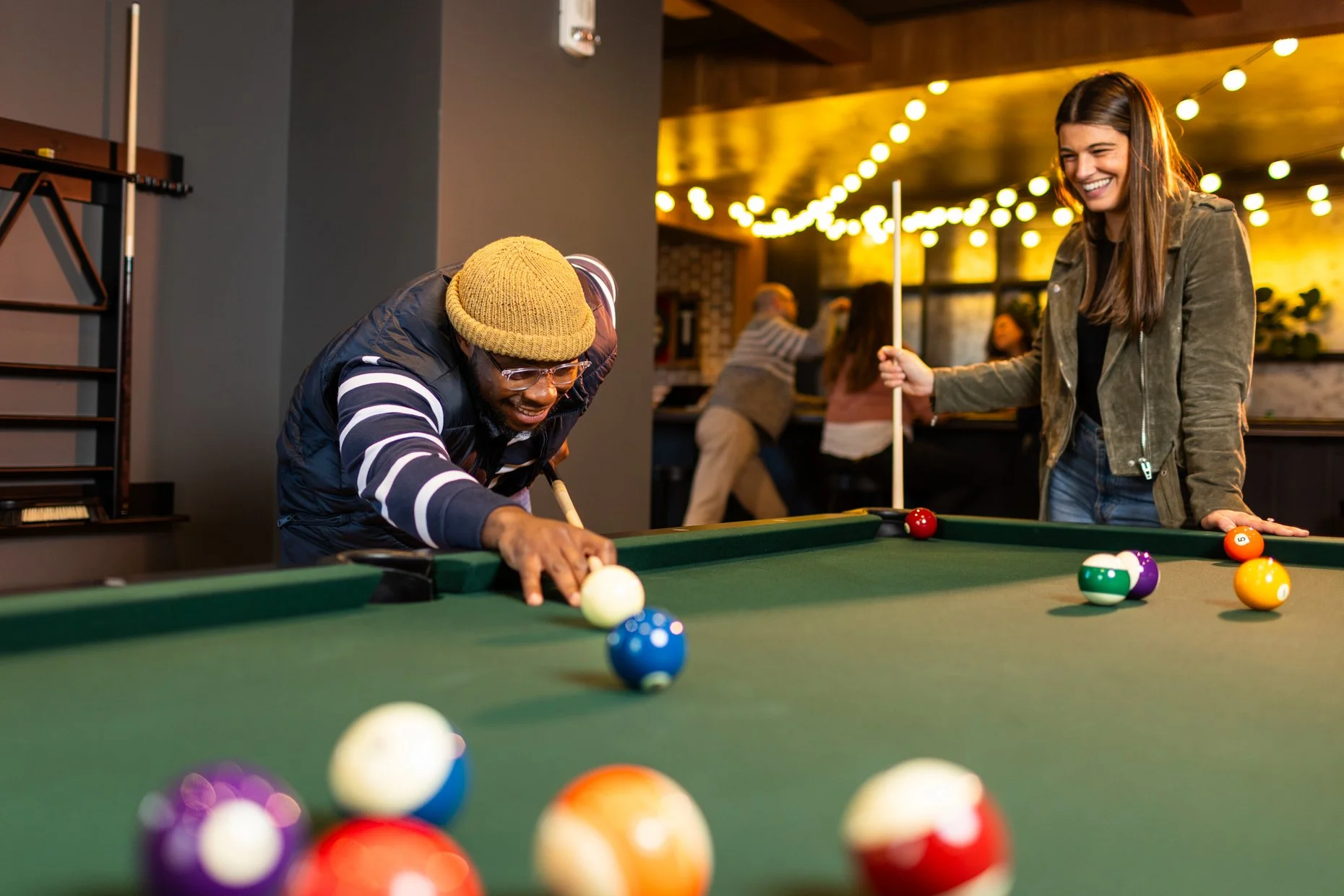 A man and a woman are playing pool at a bar or pool hall. The man is aiming to hit the cue ball, smiling, wearing glasses, a striped shirt, a black vest, and a yellow beanie. The woman is holding a cue stick, smiling, with long brown hair, wearing a green jacket and blue jeans. The pool table has several balls, including the striped and solid balls, with a background of other patrons and warm yellow string lights.
