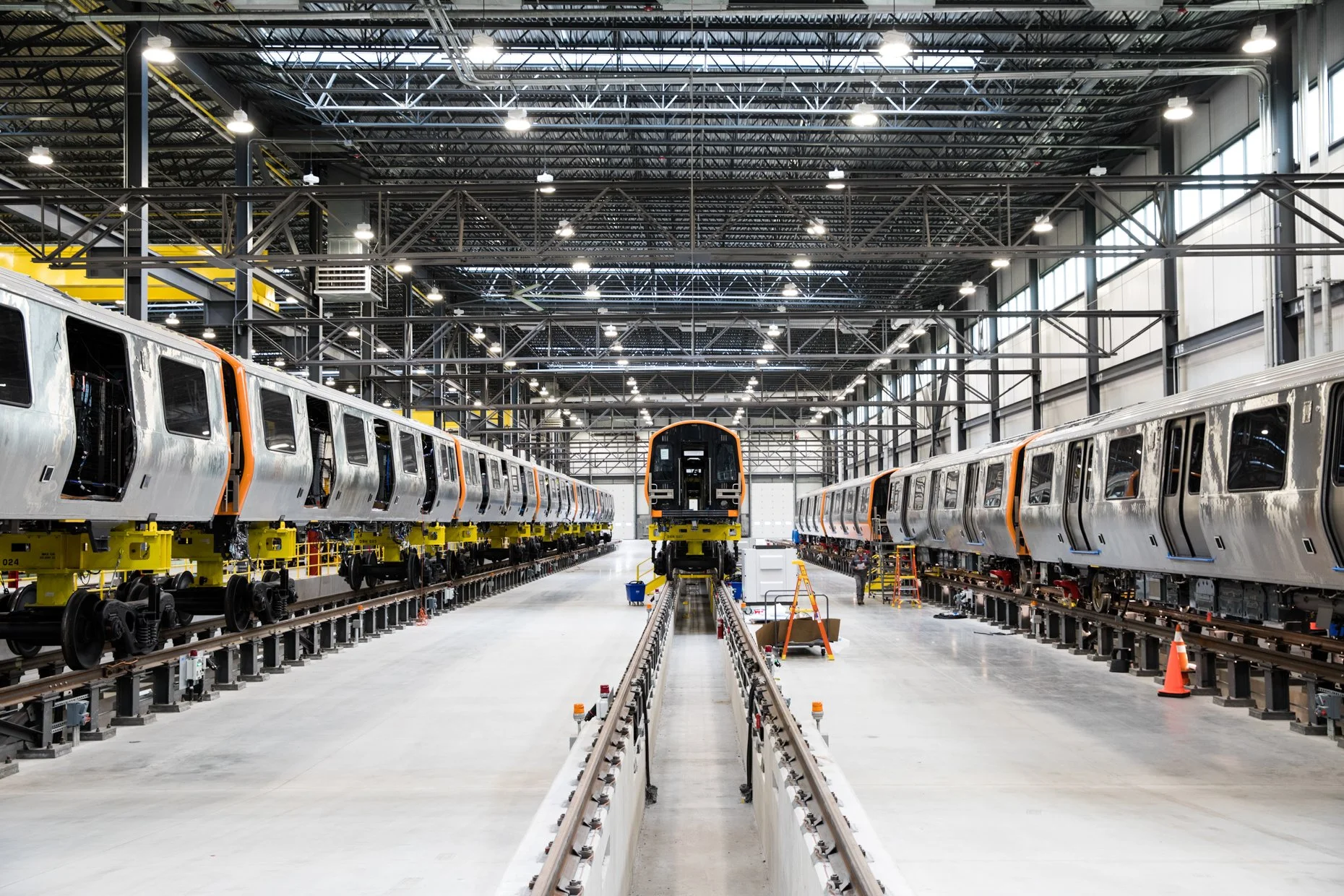 Inside a train manufacturing factory, two partially assembled trains are on tracks, with tools and equipment surrounding them.
