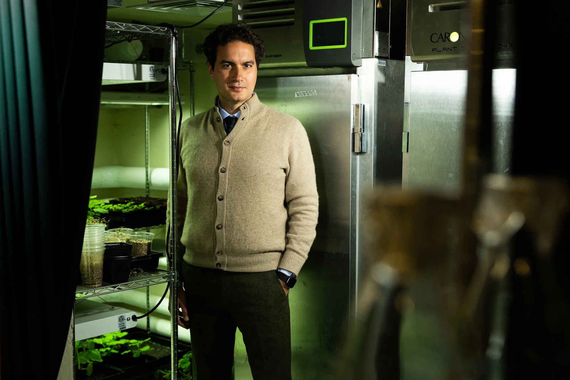 A man standing in a laboratory or indoor farm area with plants and seedlings on metal shelves, surrounded by stainless steel equipment.