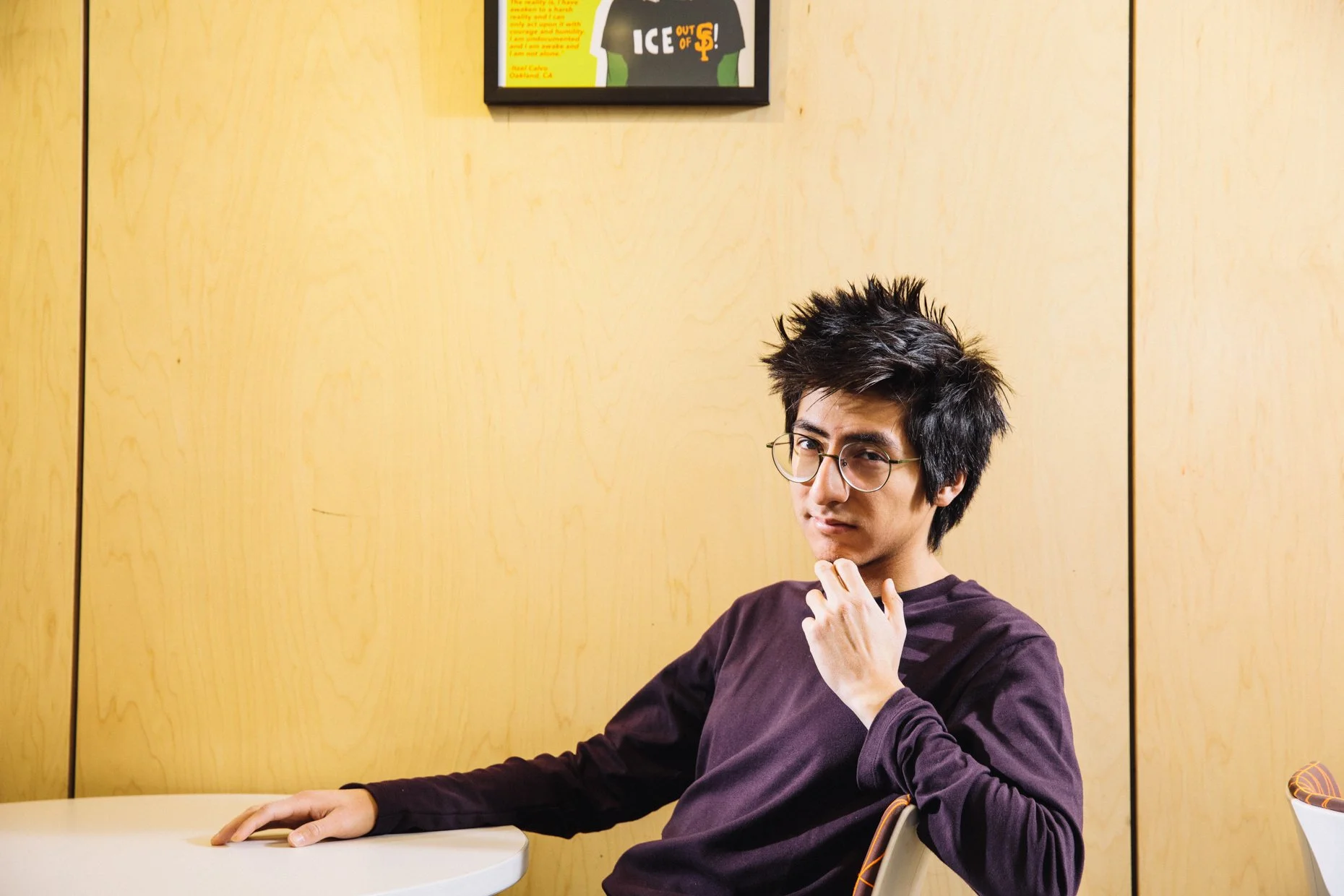 A young man with glasses and spiky black hair sitting at table in front of a light wood wall, with a contemplative expression, wearing a dark maroon shirt.