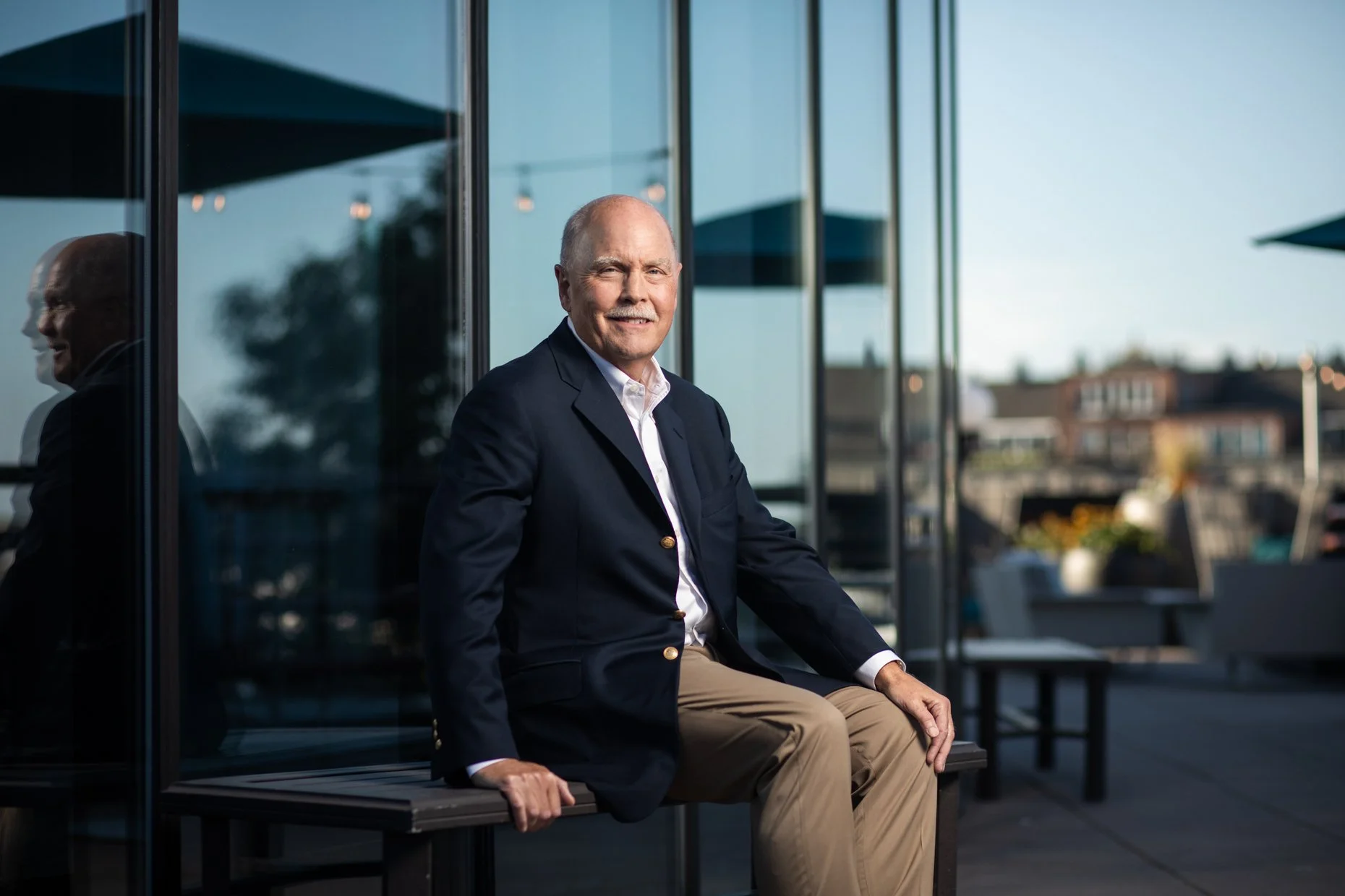 An elderly man in a navy blazer and khaki pants sitting on a bench outside a modern building, smiling at the camera, with cityscape in the background and reflections on the glass windows.