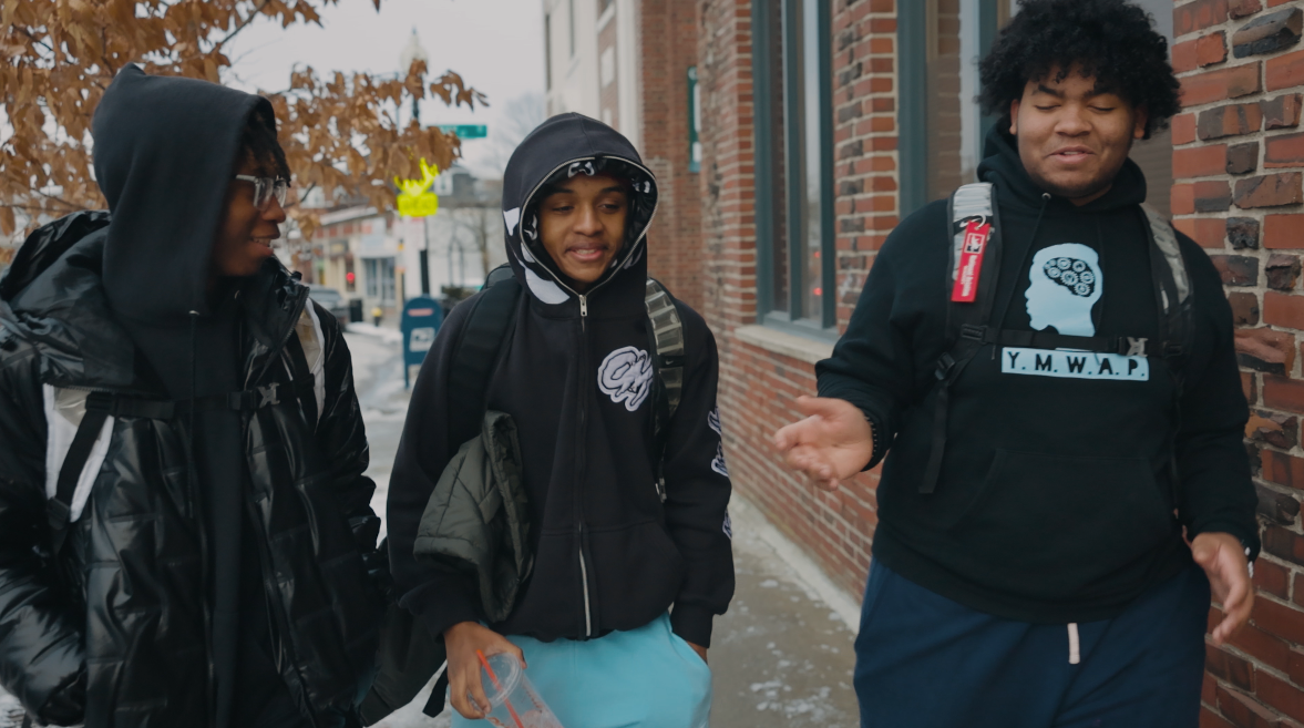 Three teenagers walking outside, talking, wearing hoodies and backpacks, near a brick building, with autumn leaves in the background.