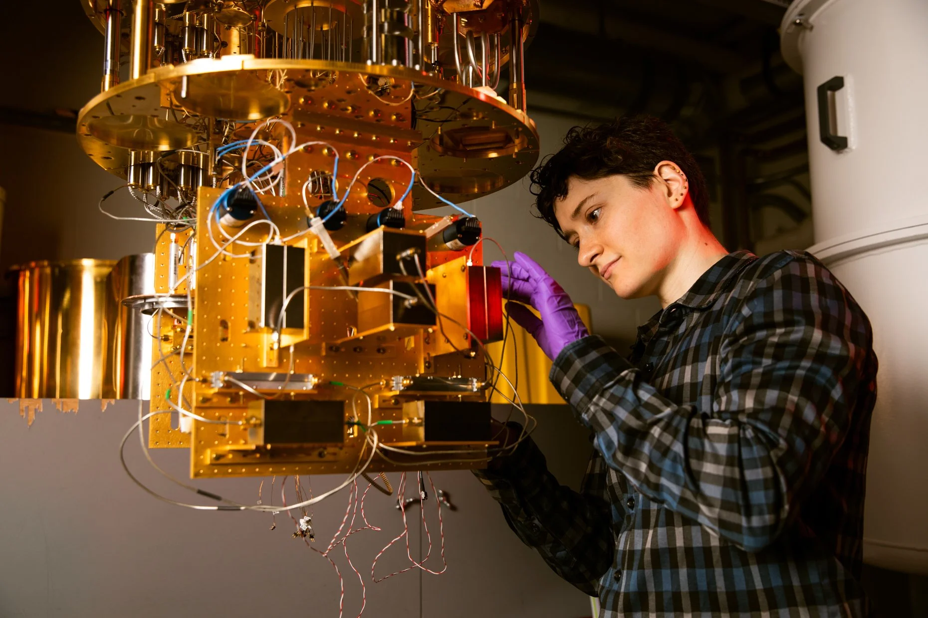 A young woman with short dark hair wearing a plaid shirt and purple gloves is working on a complex piece of scientific equipment with wires and metallic components in a lab setting.