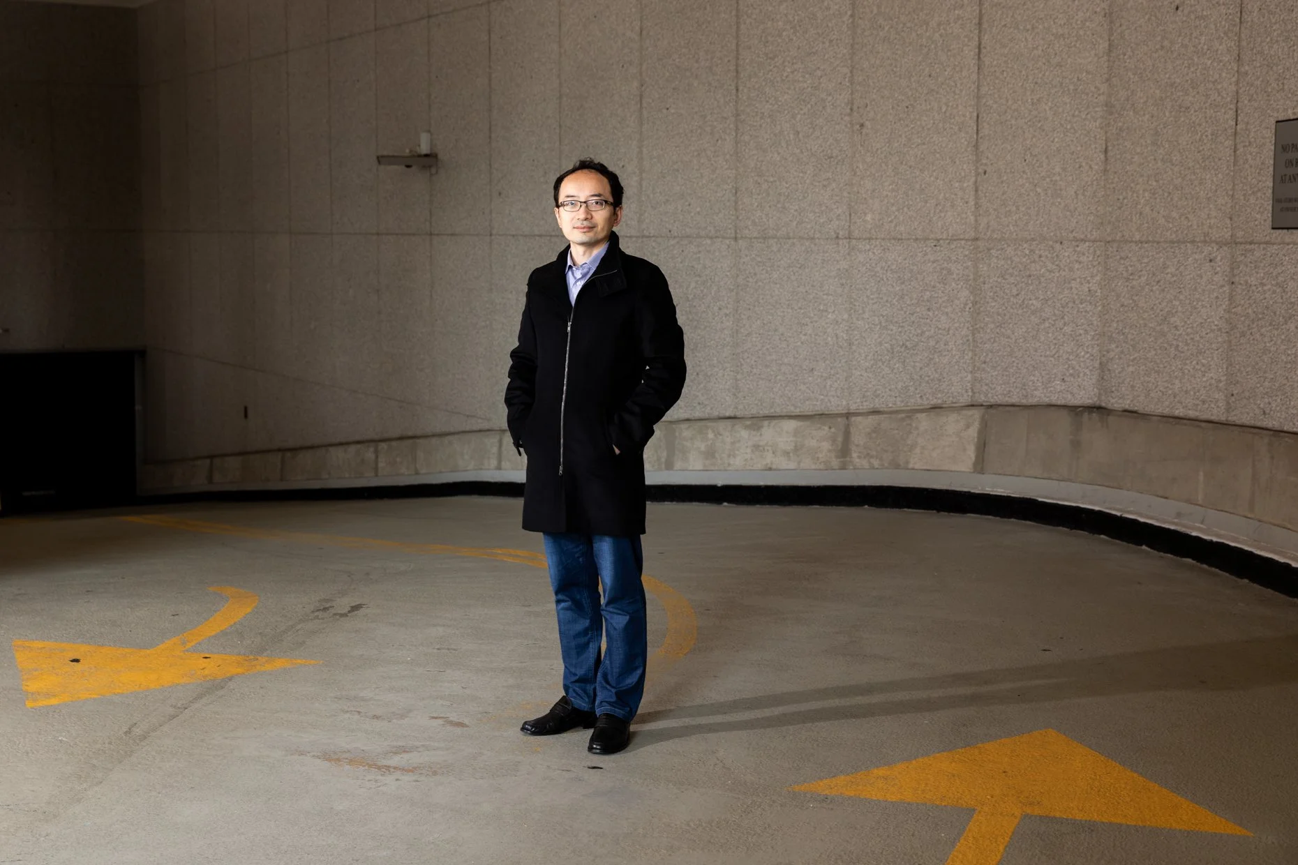A man standing in an empty parking garage with one hand in his coat pocket. He is wearing glasses, a black coat, blue jeans, and black shoes. The background features a plain concrete wall and a curved yellow parking guide line on the ground.