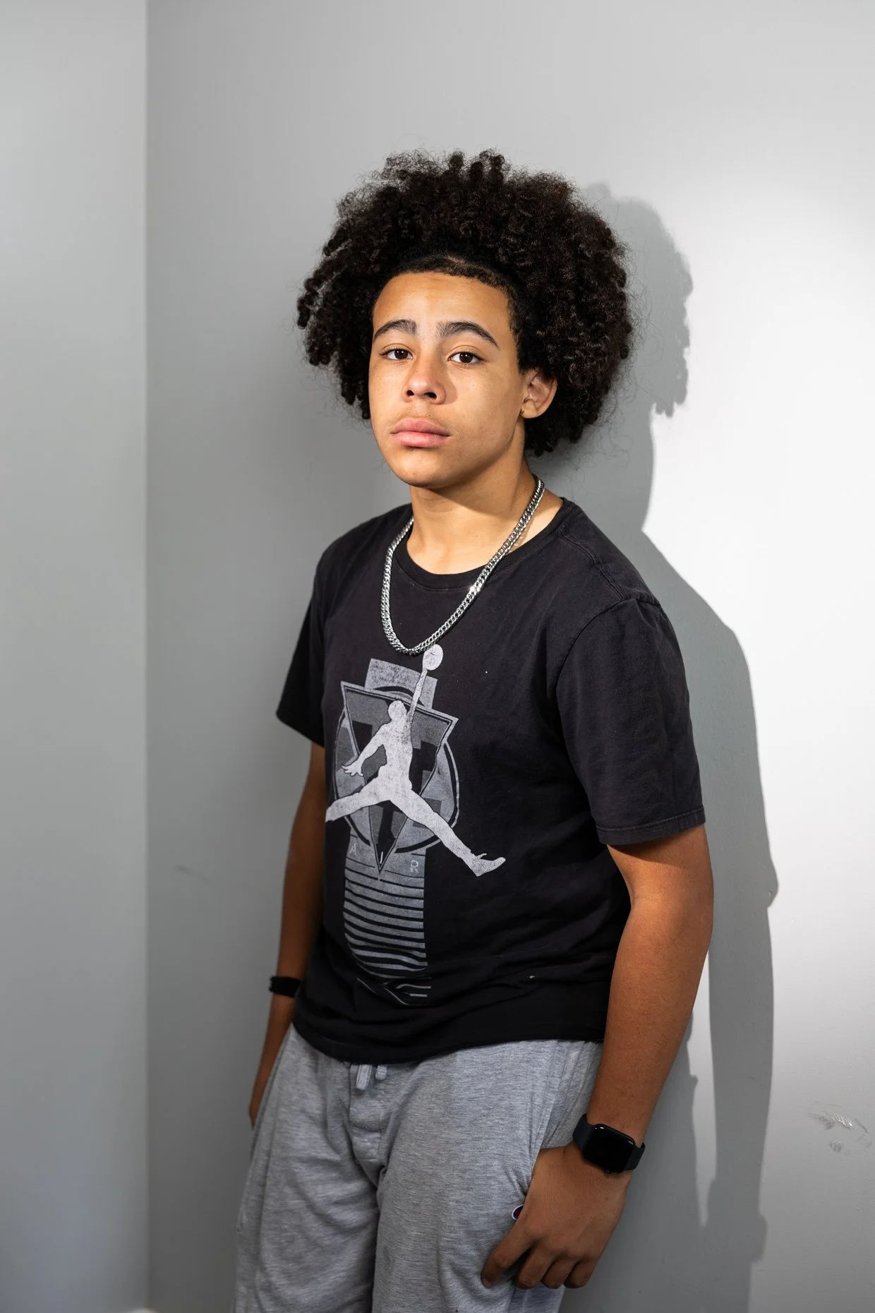 Young man with curly hair standing against a white wall, wearing a black T-shirt with a graphic design, a silver chain, a black wristband, and gray sweatpants.