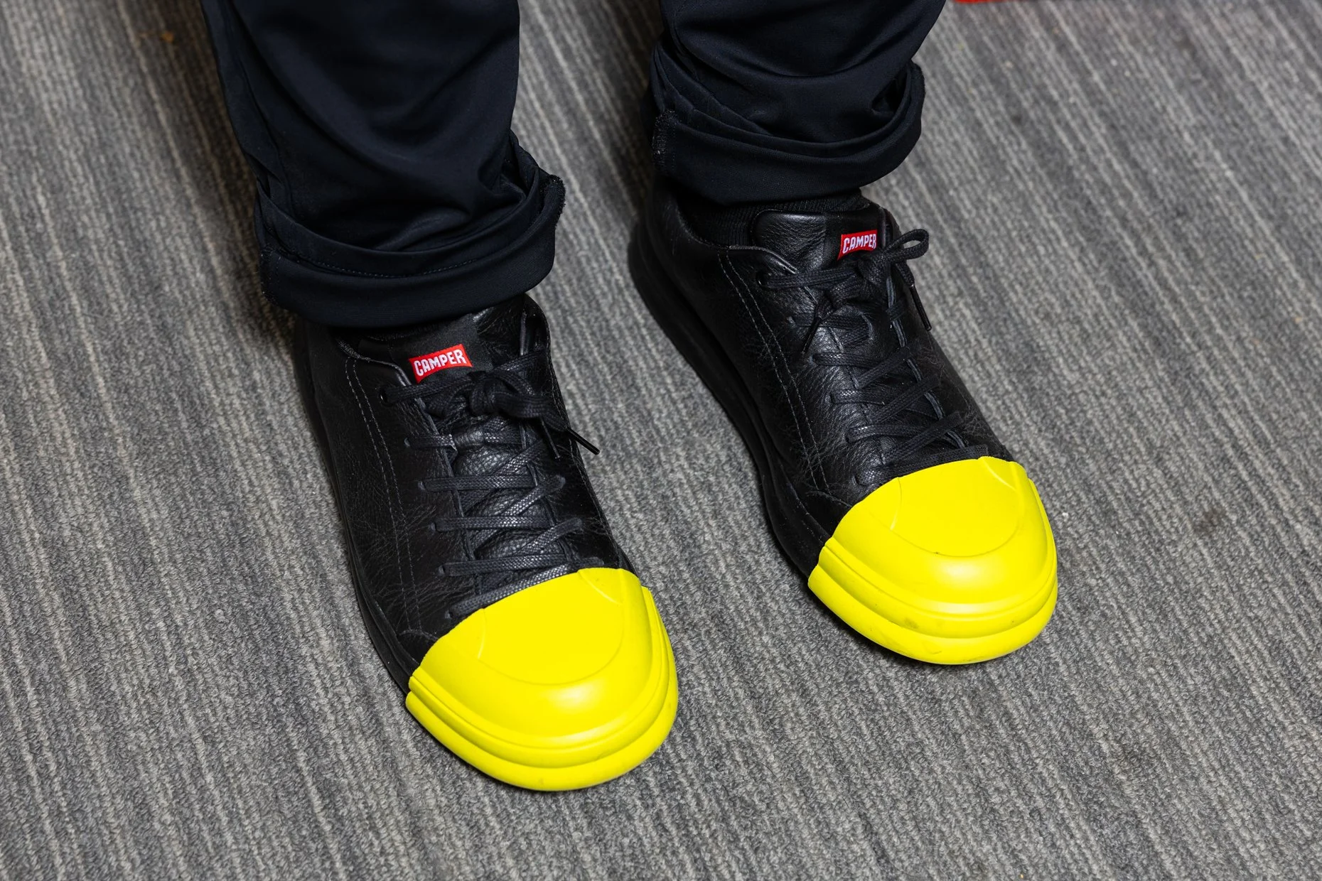 Black safety boots with yellow toe caps, worn with black pants, on a gray textured floor.