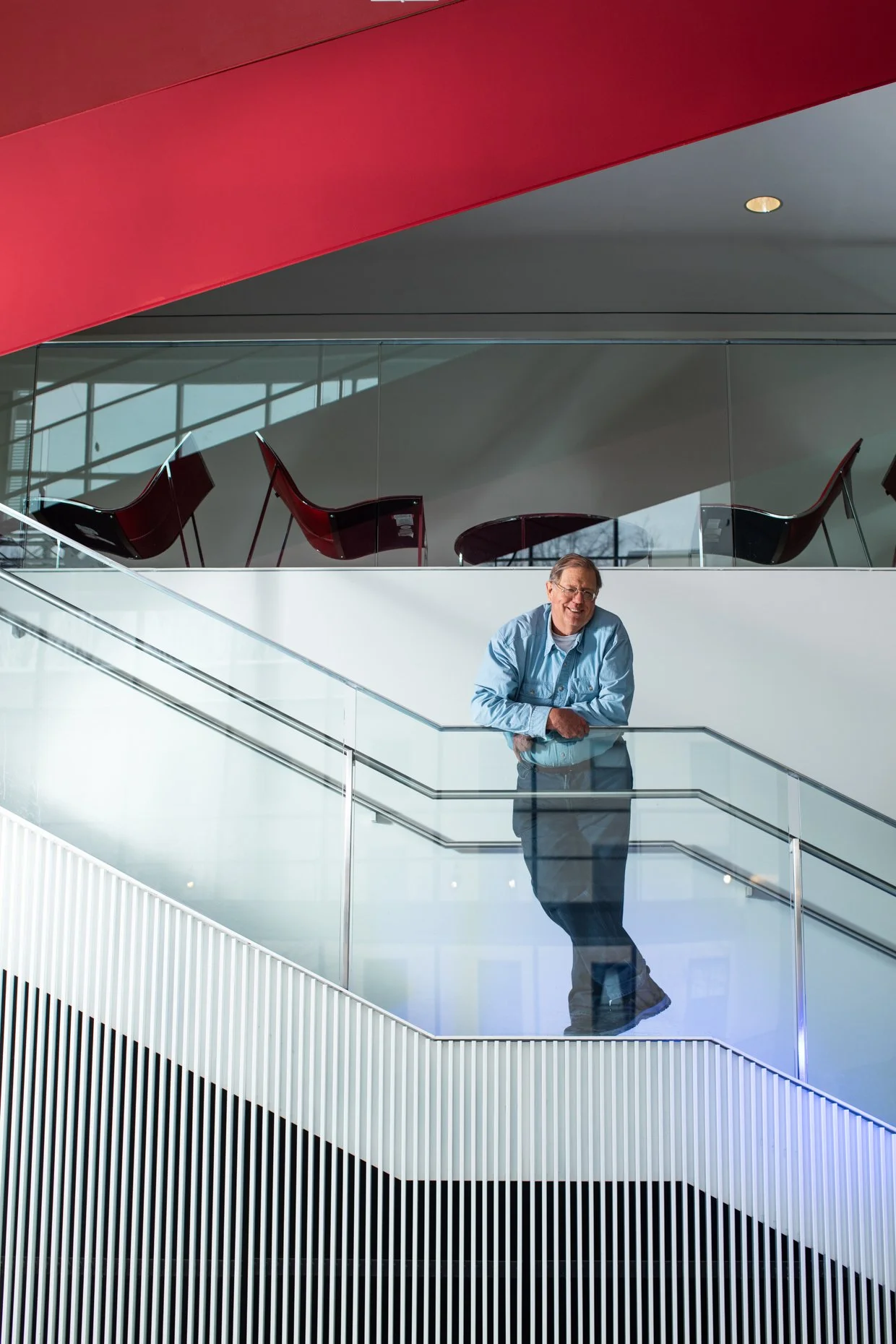A man with glasses and a blue shirt leaning on a glass railing inside a modern building with white and black interior design, and red, black, and glass furnishings.