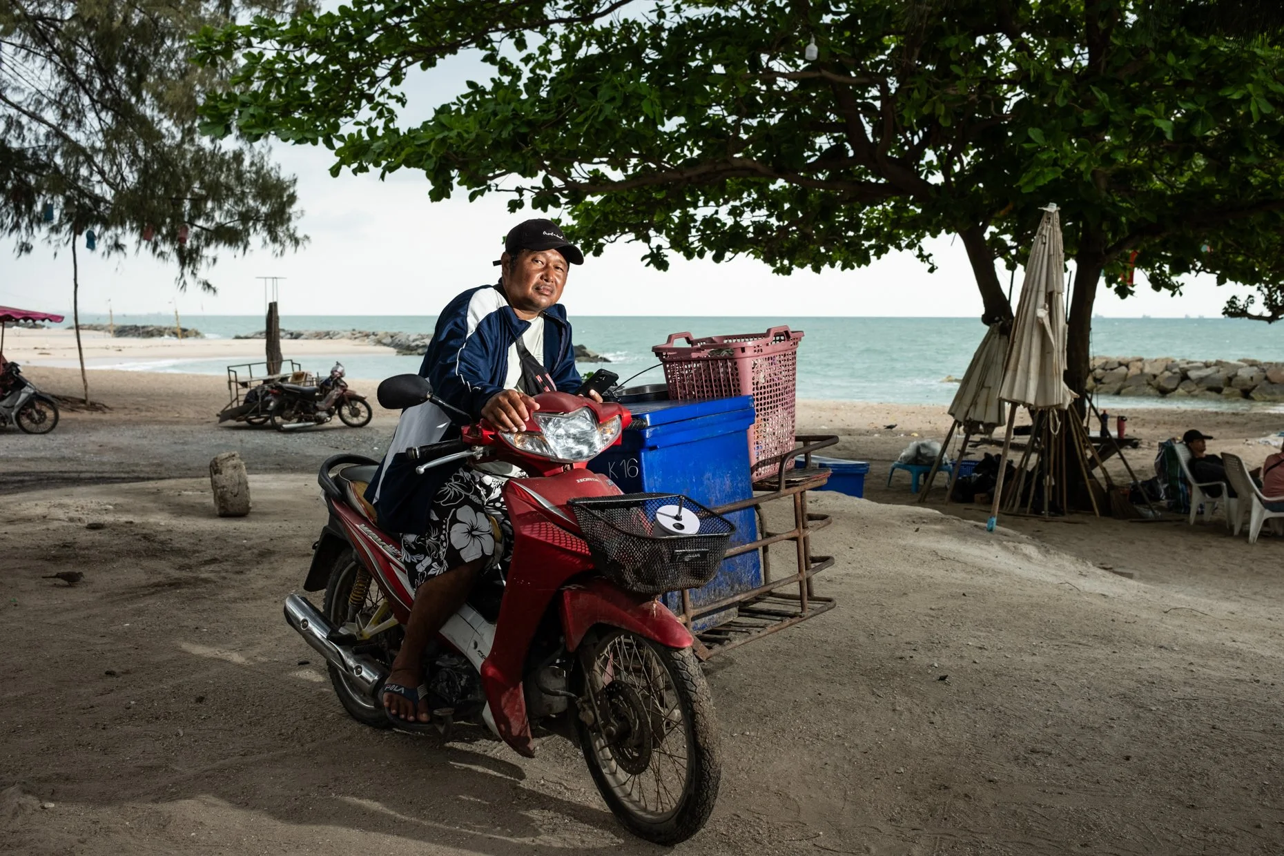 A man sitting on a red motorbike at a beachside setup, with trees providing shade and a view of the ocean in the background. There are motorcycles, shopping carts, and beach umbrellas around.