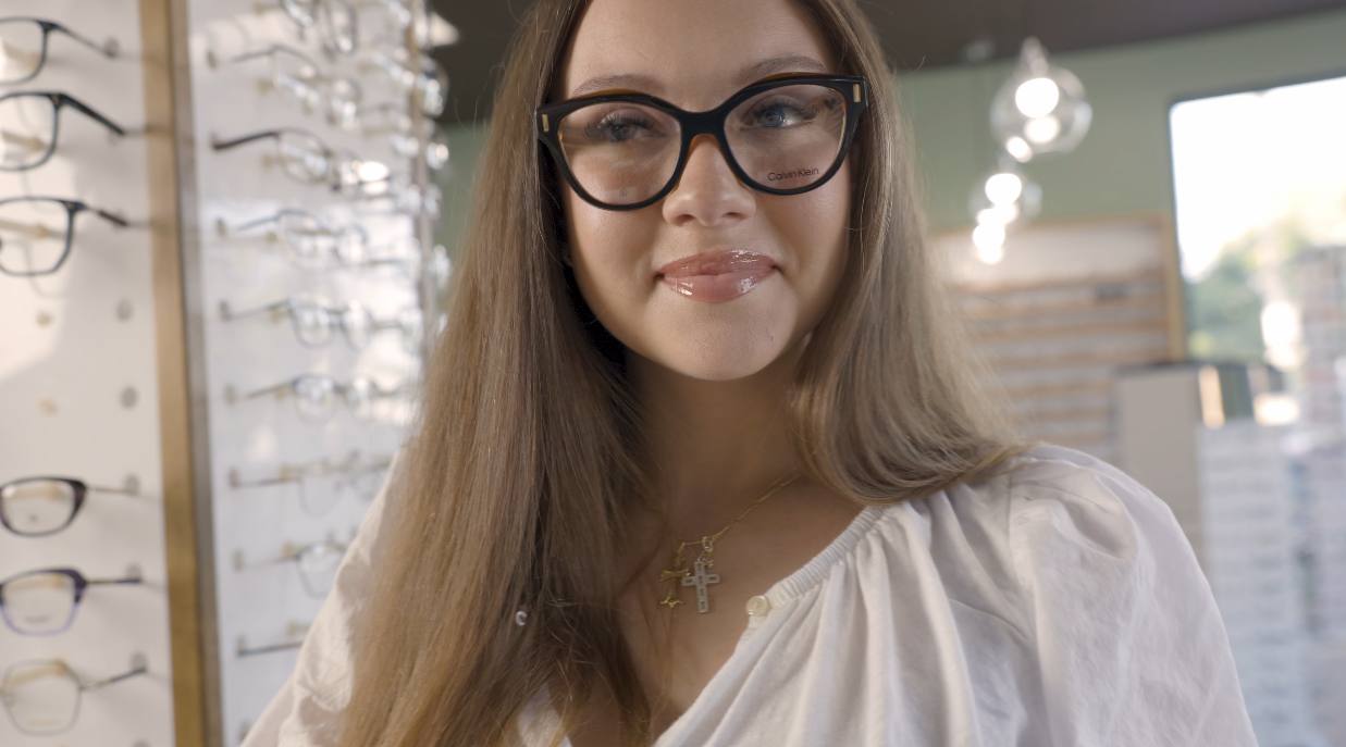 A woman with long brown hair wearing black glasses and a white blouse, standing in an eyewear store with multiple eyeglasses displayed on a wall behind her.
