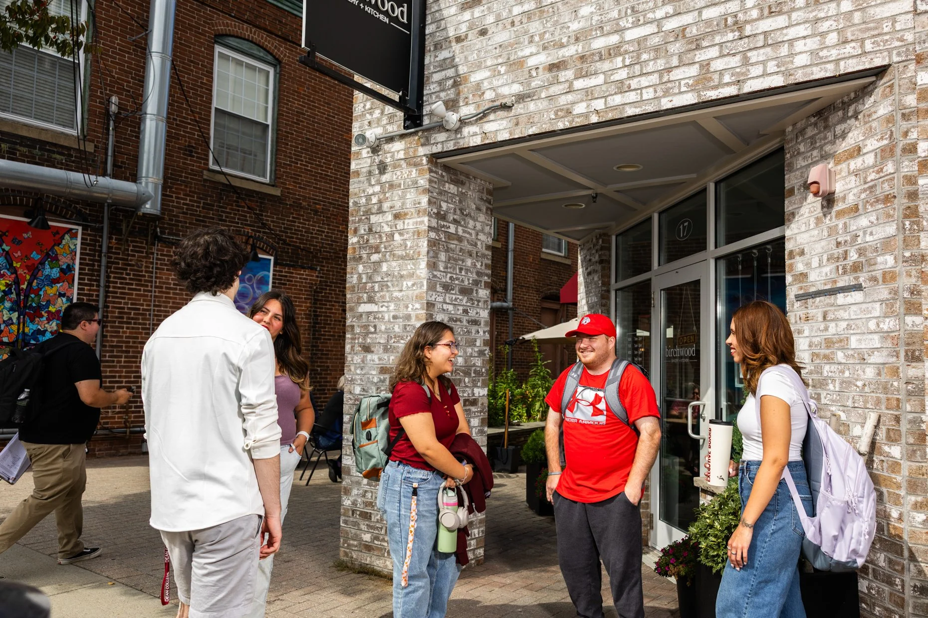 A group of five young adults standing outside a brick building, engaged in conversation, with some smiling and carrying backpacks, in an urban setting.