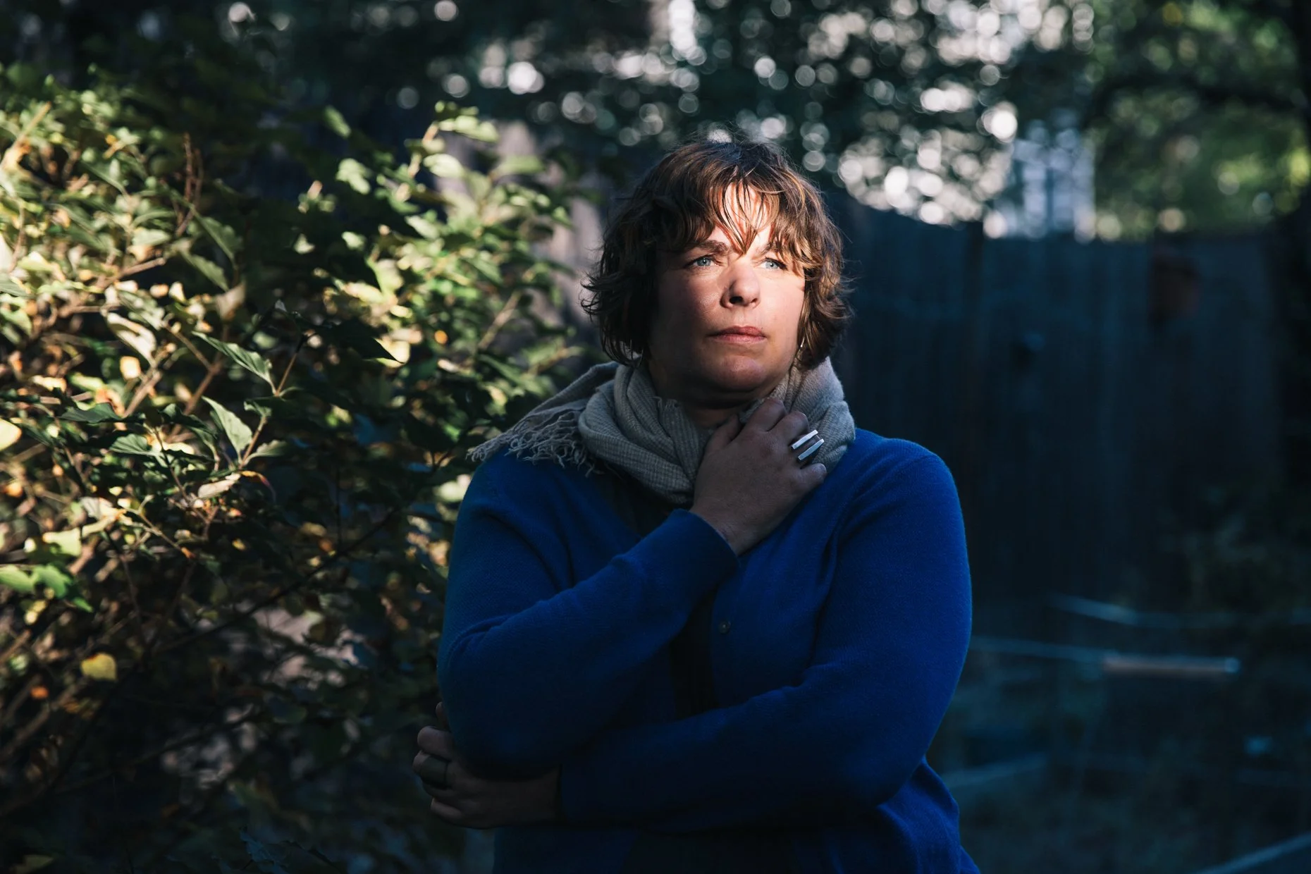 A woman with short, wavy brown hair and blue eyes stands outdoors near bushes and trees, wearing a blue sweater and a gray scarf. She is holding her throat with one hand and has a serious expression, with sunlight filtering through the trees in the background.