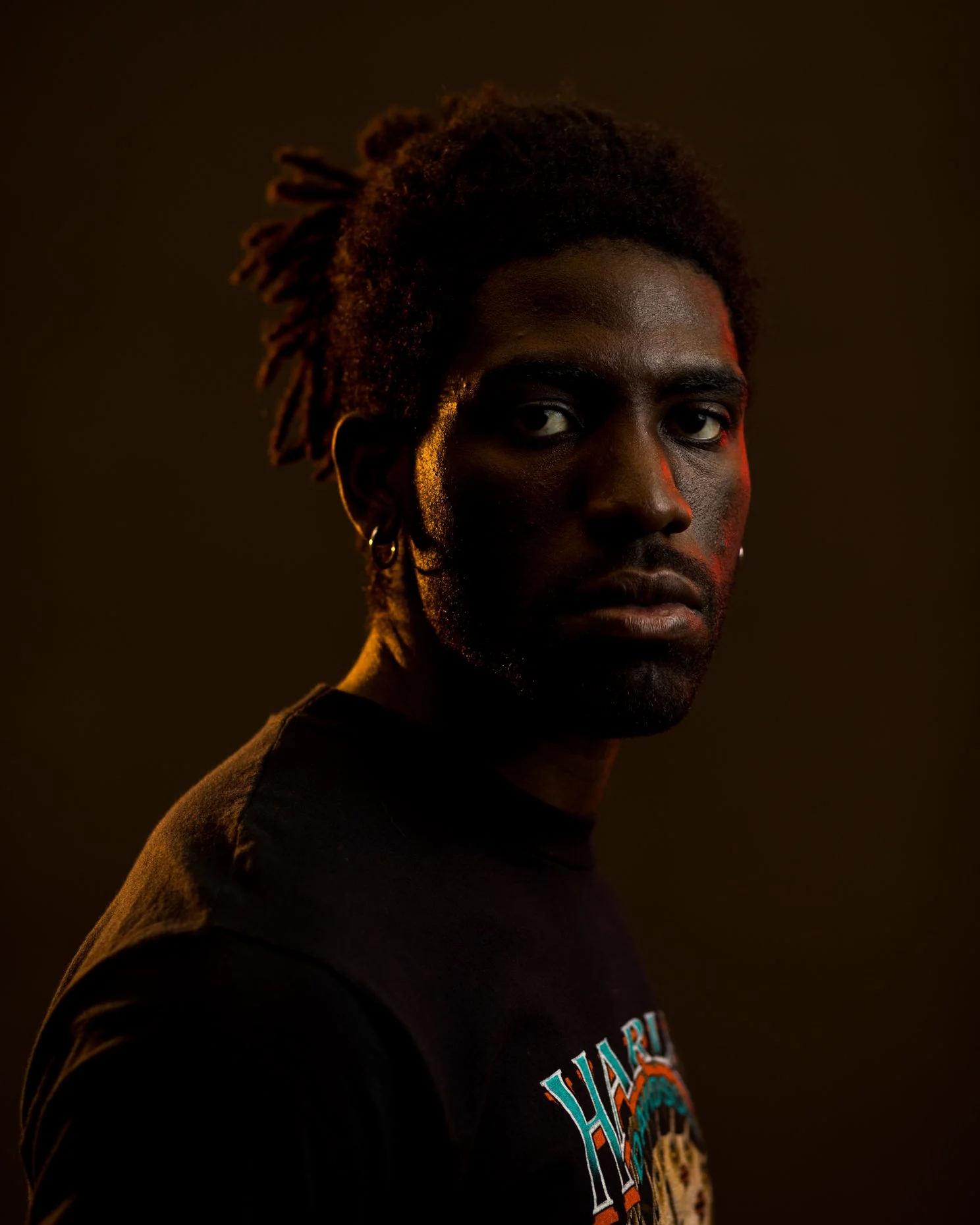 Close-up portrait of a Black man with dreadlocks, wearing a black shirt with colorful text, in a dark background with subtle warm lighting.