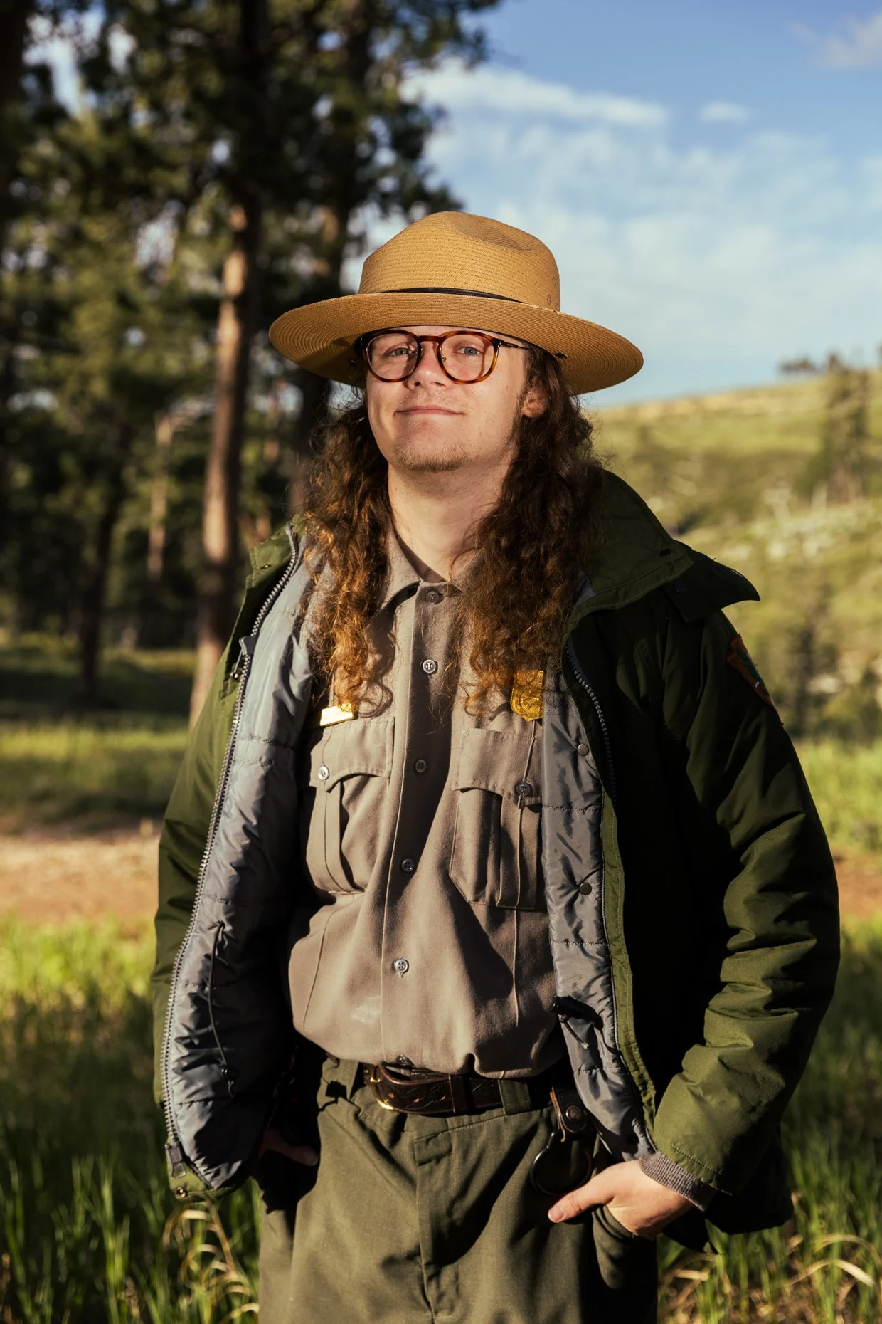 A man with long curly hair and glasses standing outdoors in a nature setting, wearing a wide-brimmed hat and a layered jacket over a beige shirt, with trees and blue sky in the background.