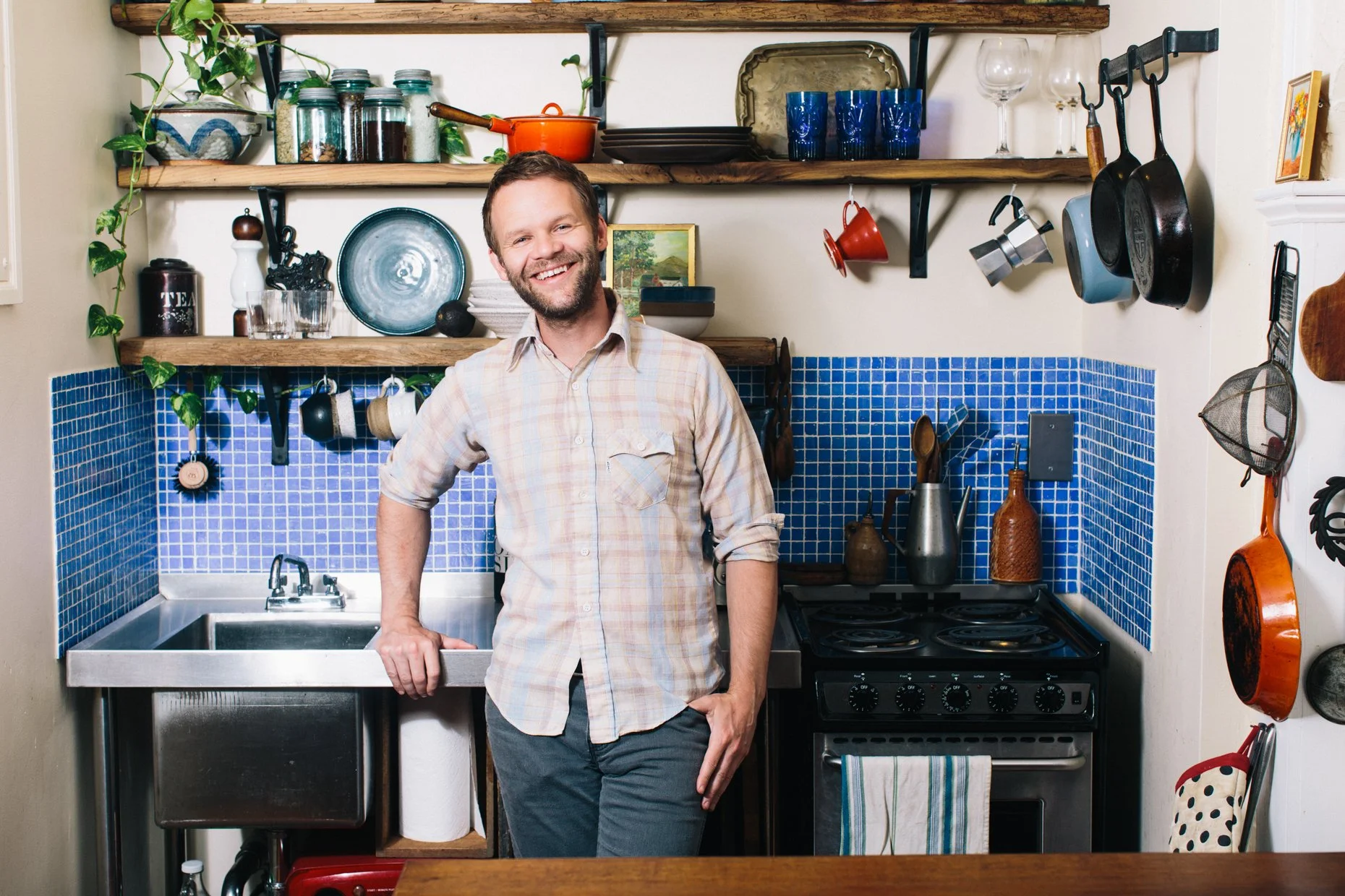 A man smiling in a kitchen with open shelving, pots, glasses, and decorative items, standing near a sink and stove.