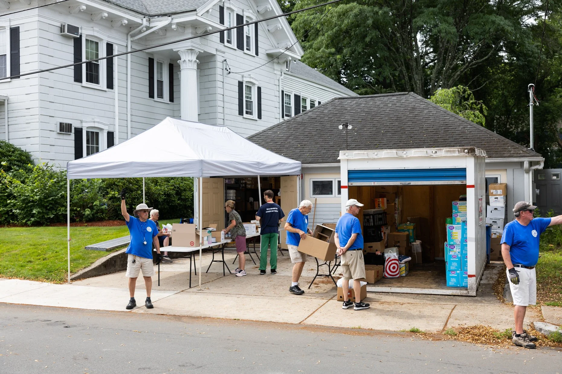 People unloading boxes into a truck and setting up a temporary outdoor event or distribution area in front of a white house with black shutters