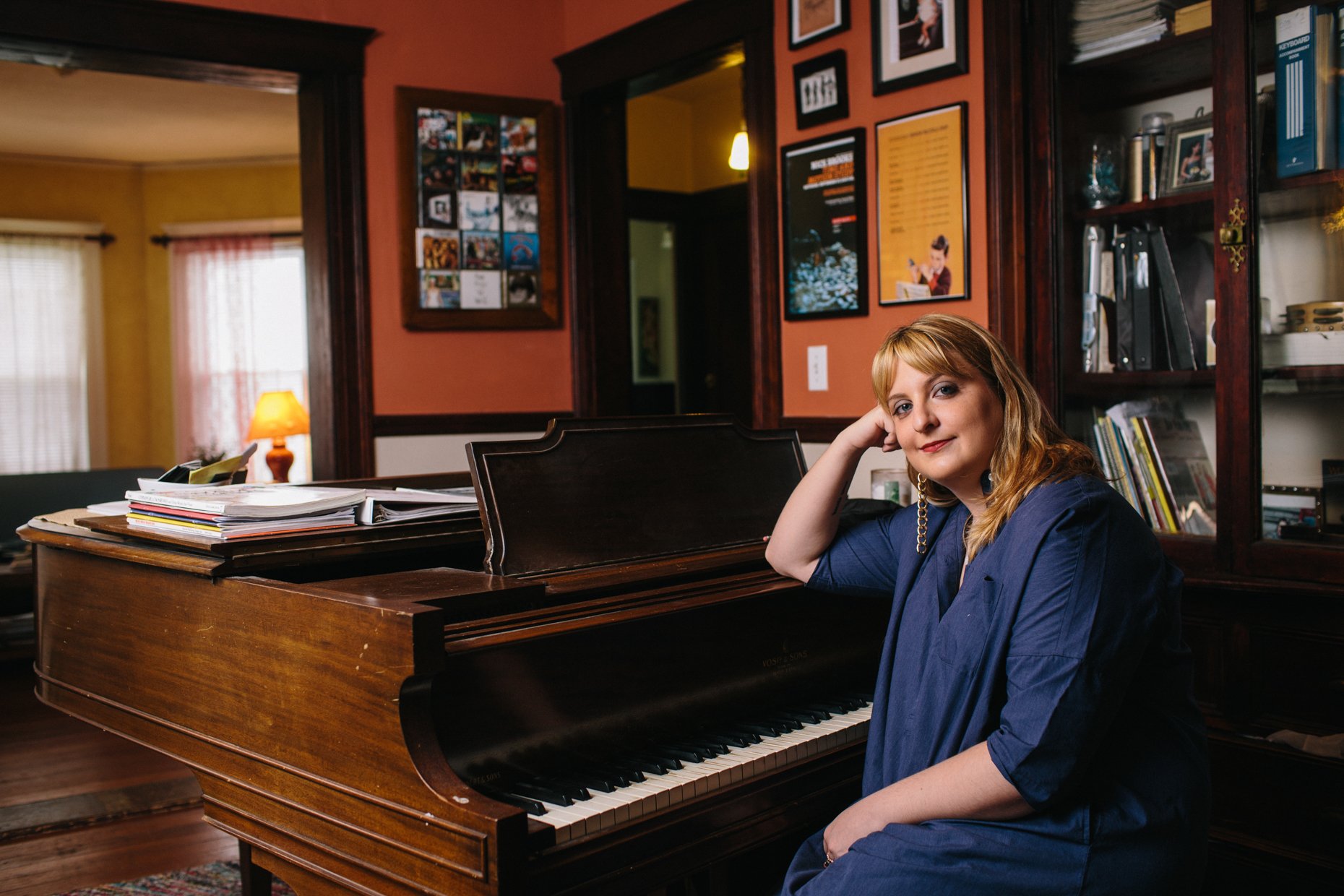 A woman with red hair and wearing a blue blazer sits at a vintage piano inside a cozy, warmly lit room with framed pictures on the walls, a shelf with books, and a window with curtains in the background.