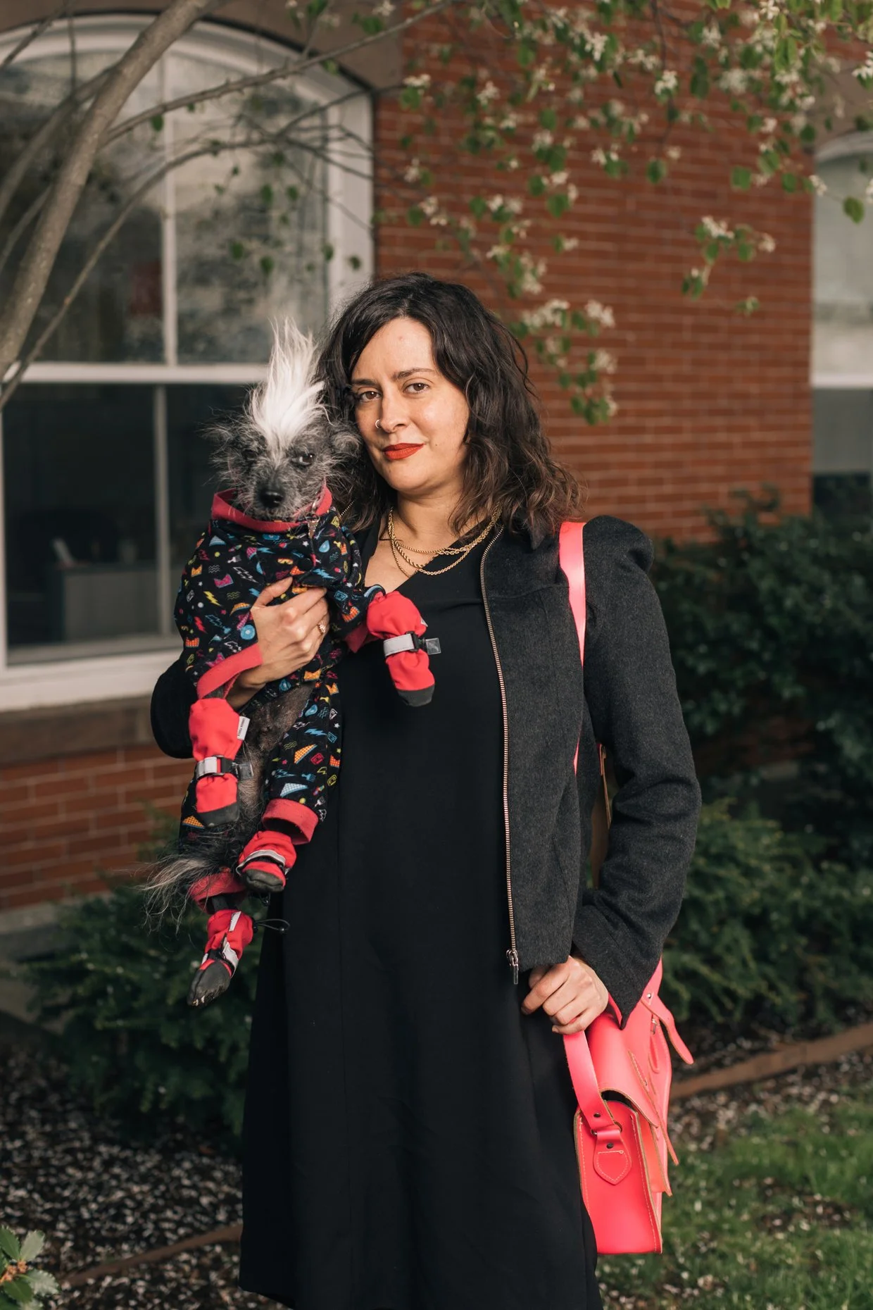 A woman with dark wavy hair and red lipstick holds a small dog with spiky black and white fur. The woman is wearing a dark coat and has a pink bag on her shoulder. They are outside in front of a red brick building with greenery and white flowers in the background.