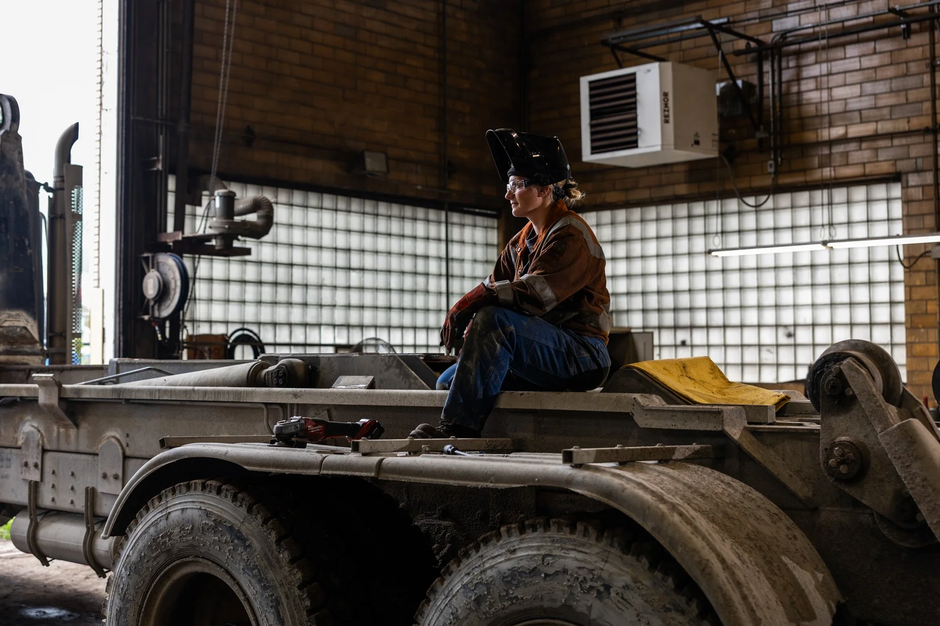 A woman wearing safety gear, including a welding helmet, sits on a construction vehicle inside an industrial building with brick walls and large glass block windows.