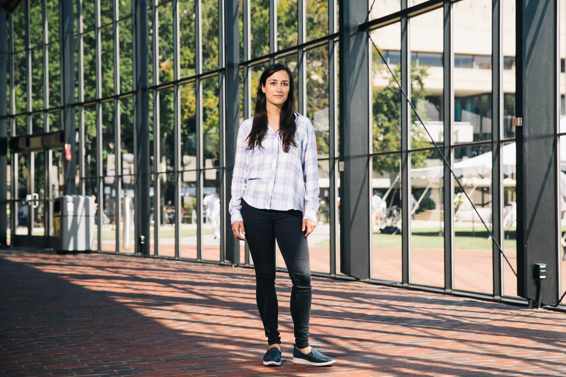 A young woman with long dark hair standing outside near a glass structure, wearing a light-colored plaid shirt, black pants, and dark slip-on shoes, with trees and a building in the background.