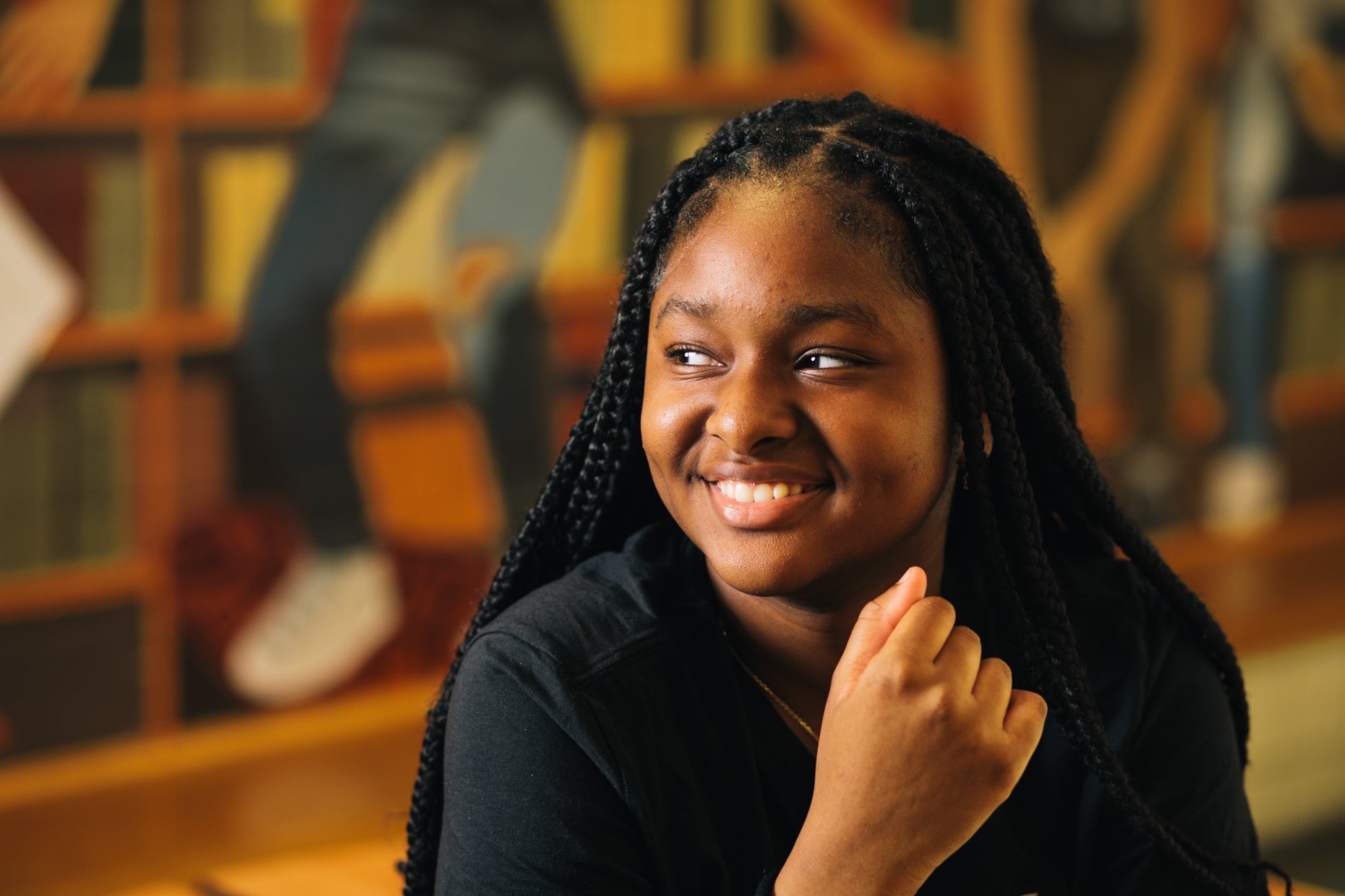 A young woman with braided hair smiling and looking to her right, sitting in a room with a background of bookshelves filled with books.