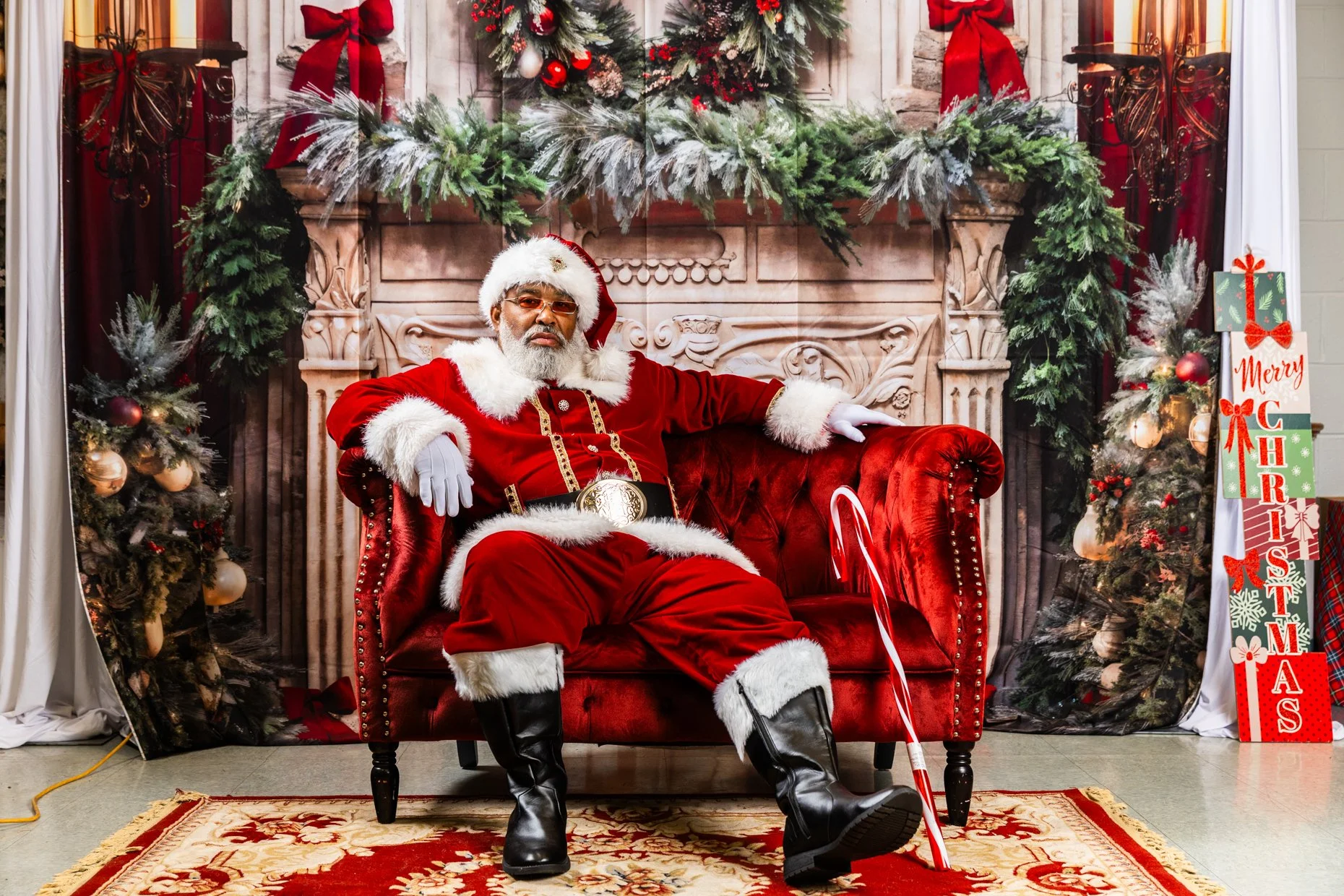 Santa Claus dressed in traditional red and white sitting on a red velvet chair with a festive Christmas background, decorated with Christmas trees, greenery, red ribbons, ornaments, and holiday signs.
