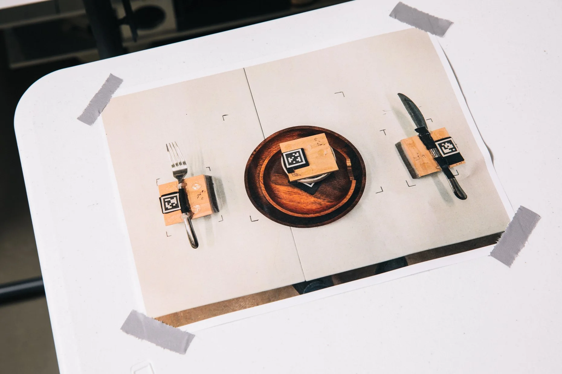 A white table with a printed photograph taped to it, depicting two slices of wood with QR code stickers on top, a fork and a knife each placed on one slice, and a small round wooden dish holding a stack of paper or cards.